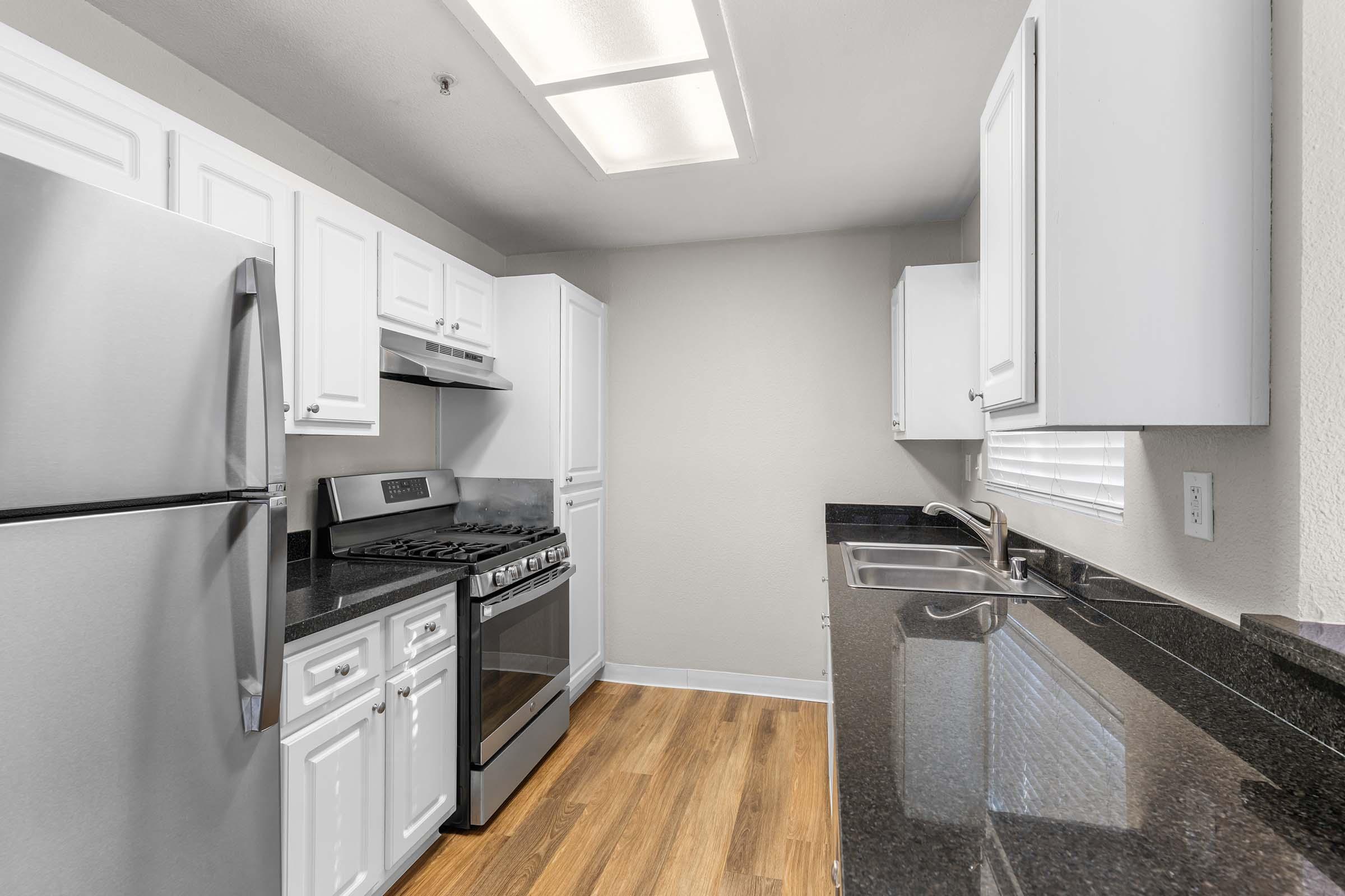 Modern kitchen featuring stainless steel appliances, including a gas range and refrigerator. It has white cabinetry, black countertops, and wood-style flooring. The room is well-lit with natural light from a window, creating a clean and contemporary atmosphere.