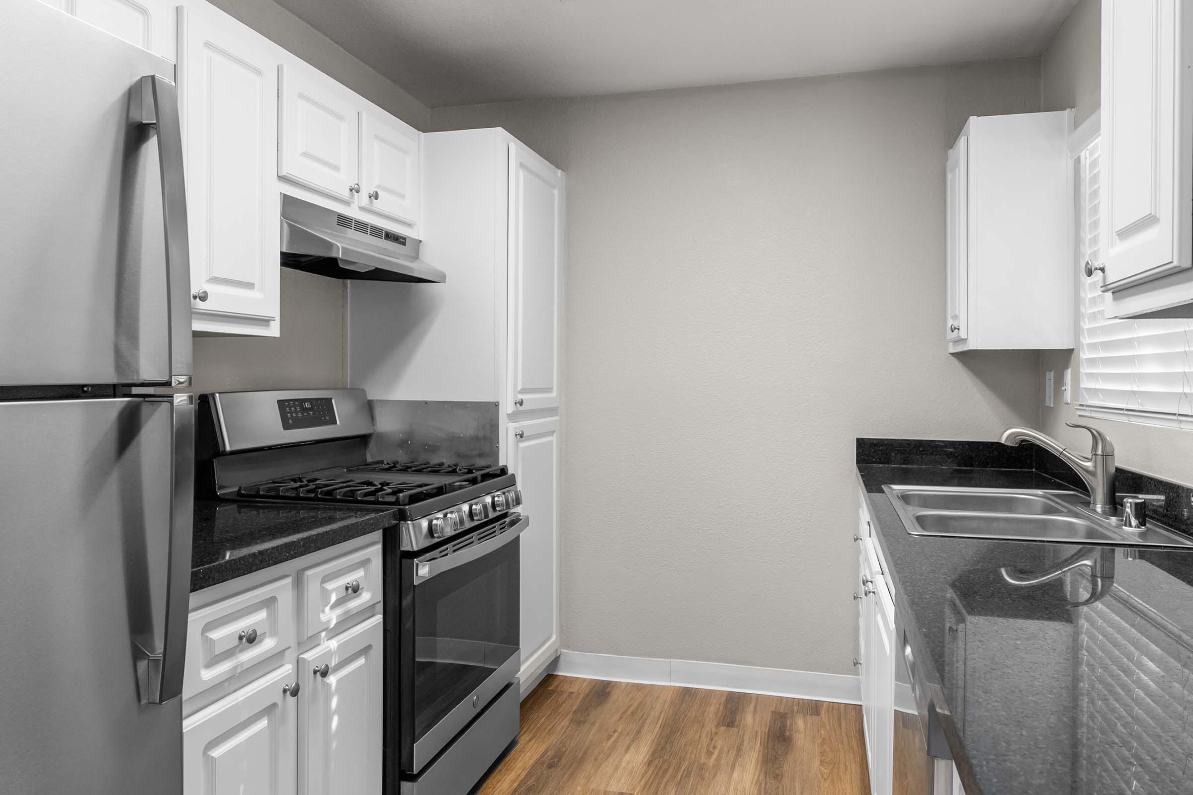Modern kitchen featuring stainless steel appliances, including a refrigerator and a gas stove. The cabinetry is white with a sleek black countertop and sink. The space has light-colored walls and wooden flooring, contributing to a clean and open feel.