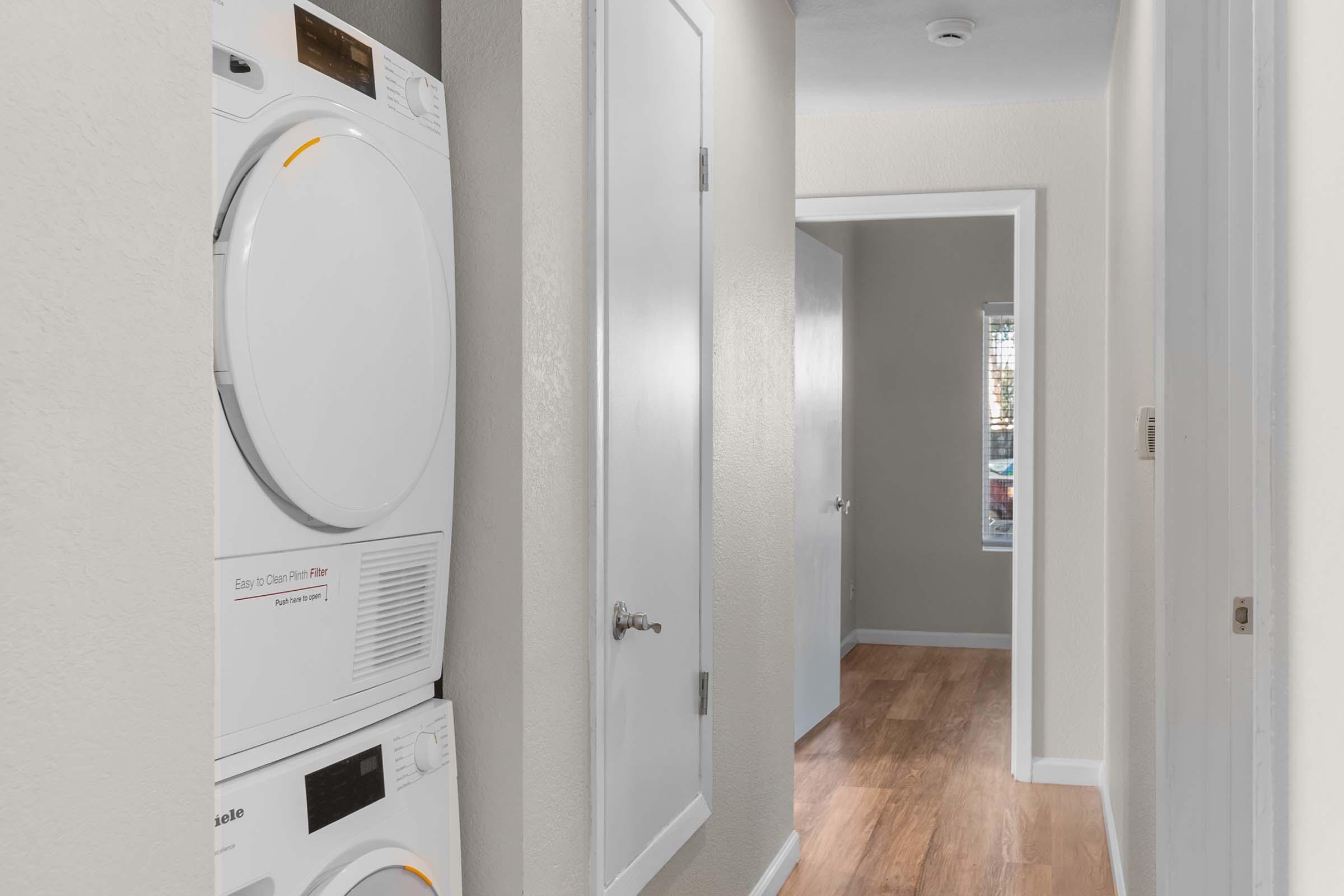A hallway featuring a stackable washer and dryer on the left, with light-colored walls and hardwood flooring. A doorway leads to another room on the right, and a window is visible at the end of the hallway, allowing natural light to enter the space.