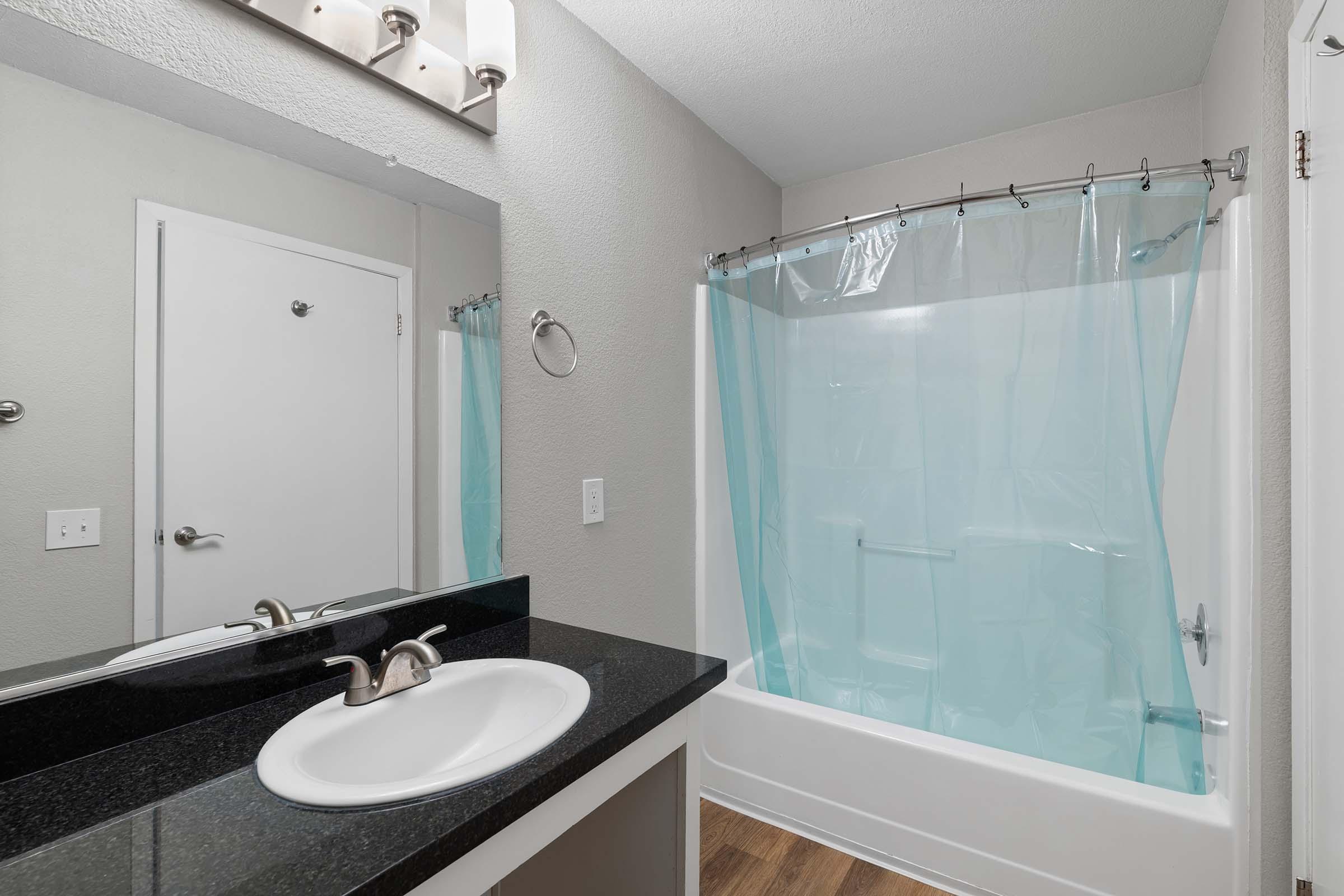 A modern bathroom featuring a sleek black countertop with a sink, a light fixture above, and a mirror. The bathtub is enclosed with a light blue shower curtain, and there is a towel ring on the wall. The floor is wood-like, adding warmth to the space.