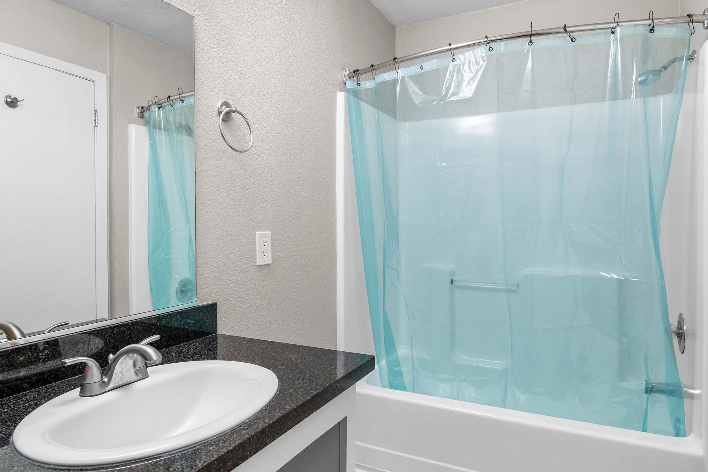 A bathroom featuring a white bathtub with a light blue shower curtain, a dark countertop with a sink, a mirror above the sink, and a towel rack on the wall. The walls are a neutral color, and there is a door partially visible, adding to the overall clean and simple aesthetic.