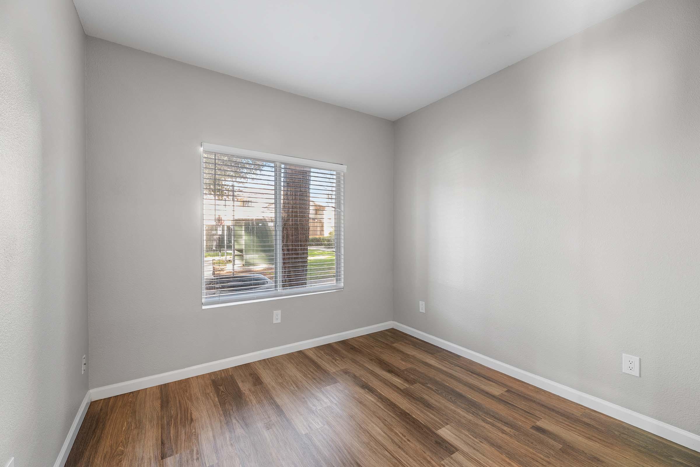 Empty room with light gray walls and hardwood flooring. A window with blinds allows light to enter, revealing a view of greenery outside. The room appears clean and well-maintained, suitable for various uses.