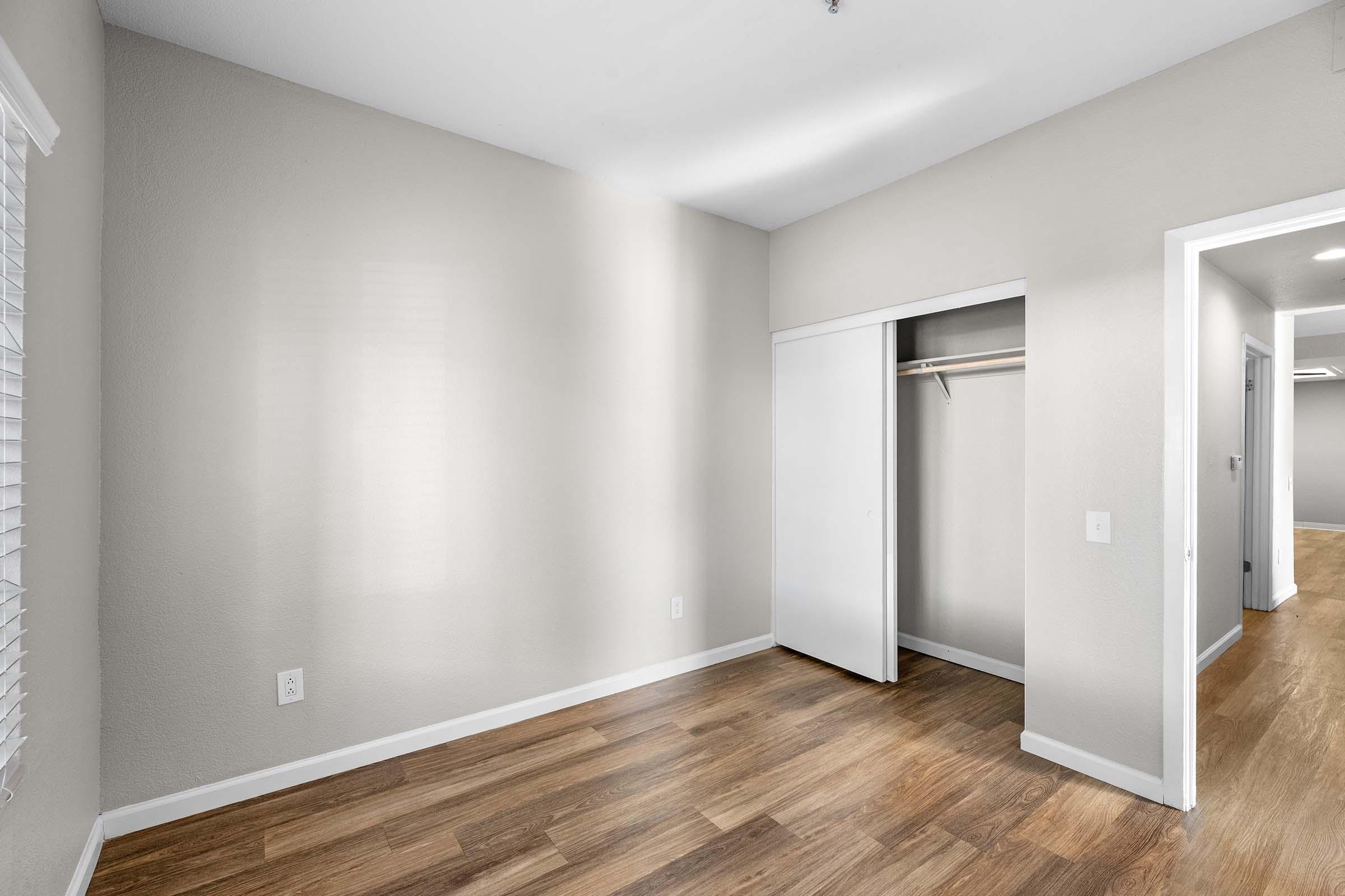 A spacious, empty room with light-colored walls and wood-like flooring. There are two doors: one leading to a closet with a sliding door and another possibly leading to a hallway. The room features natural light from a window with white blinds.