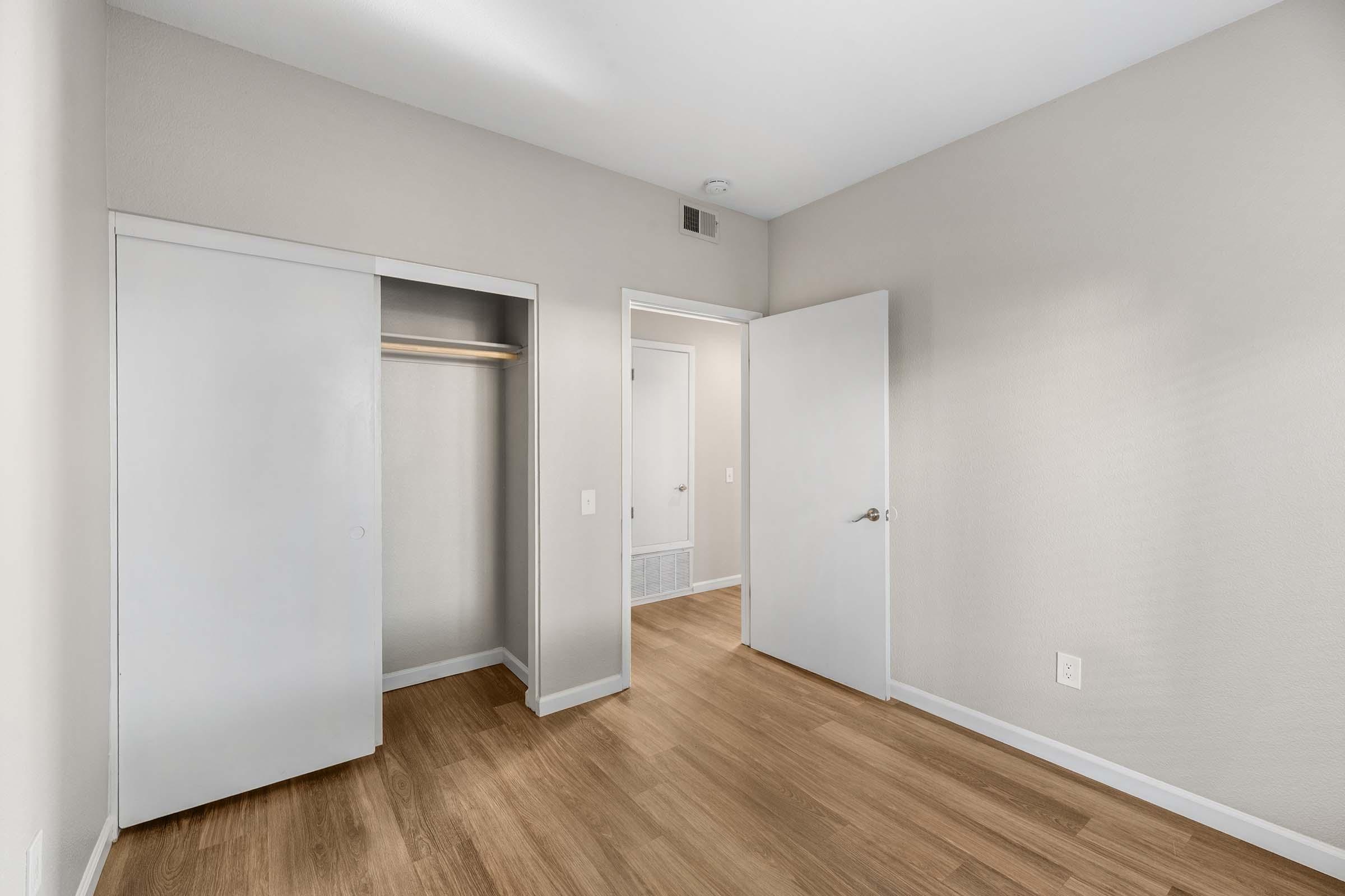 A spacious, empty room featuring light-colored walls and wooden flooring. It includes a closet with sliding doors, an open doorway leading to another room, and a simple design, creating a modern and minimalist aesthetic. Natural light is likely to illuminate the space.