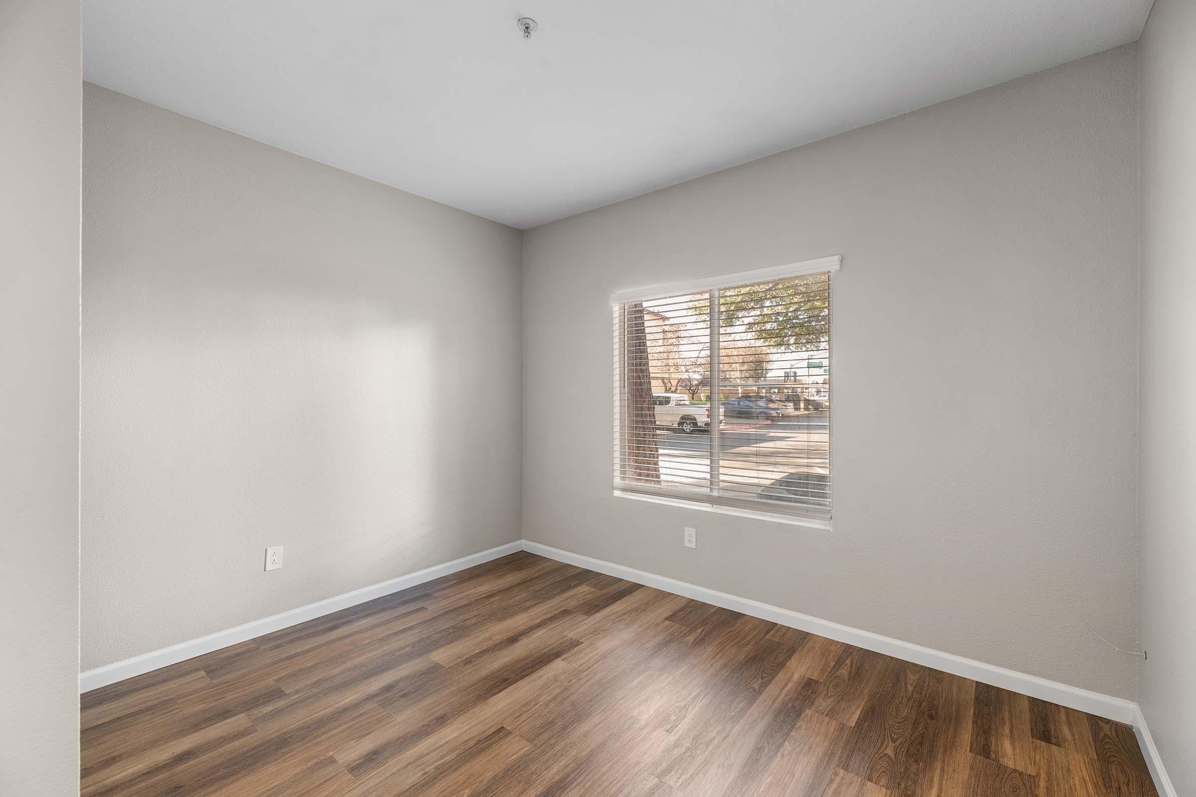Empty room with light gray walls and brown wood laminate flooring. A window with white blinds is on the right, letting in natural light and showing a view of the outside. There are no furnishings or decorations in the space, creating a clean and minimalistic appearance.