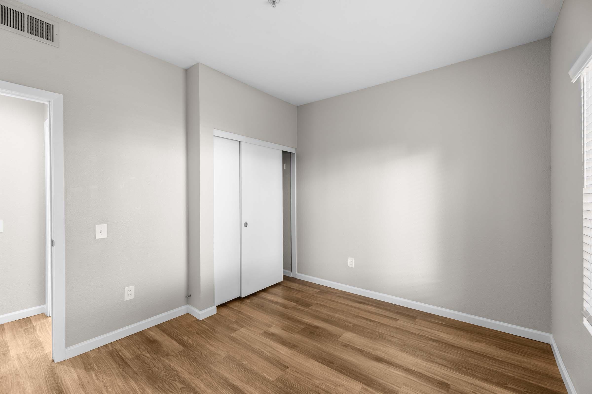 Empty room with light gray walls and wooden flooring. Two doors are visible: one leading to another room and the other a closet. A window allows natural light to enter, creating a bright and spacious feel. The room is unfurnished, emphasizing its potential for customization.
