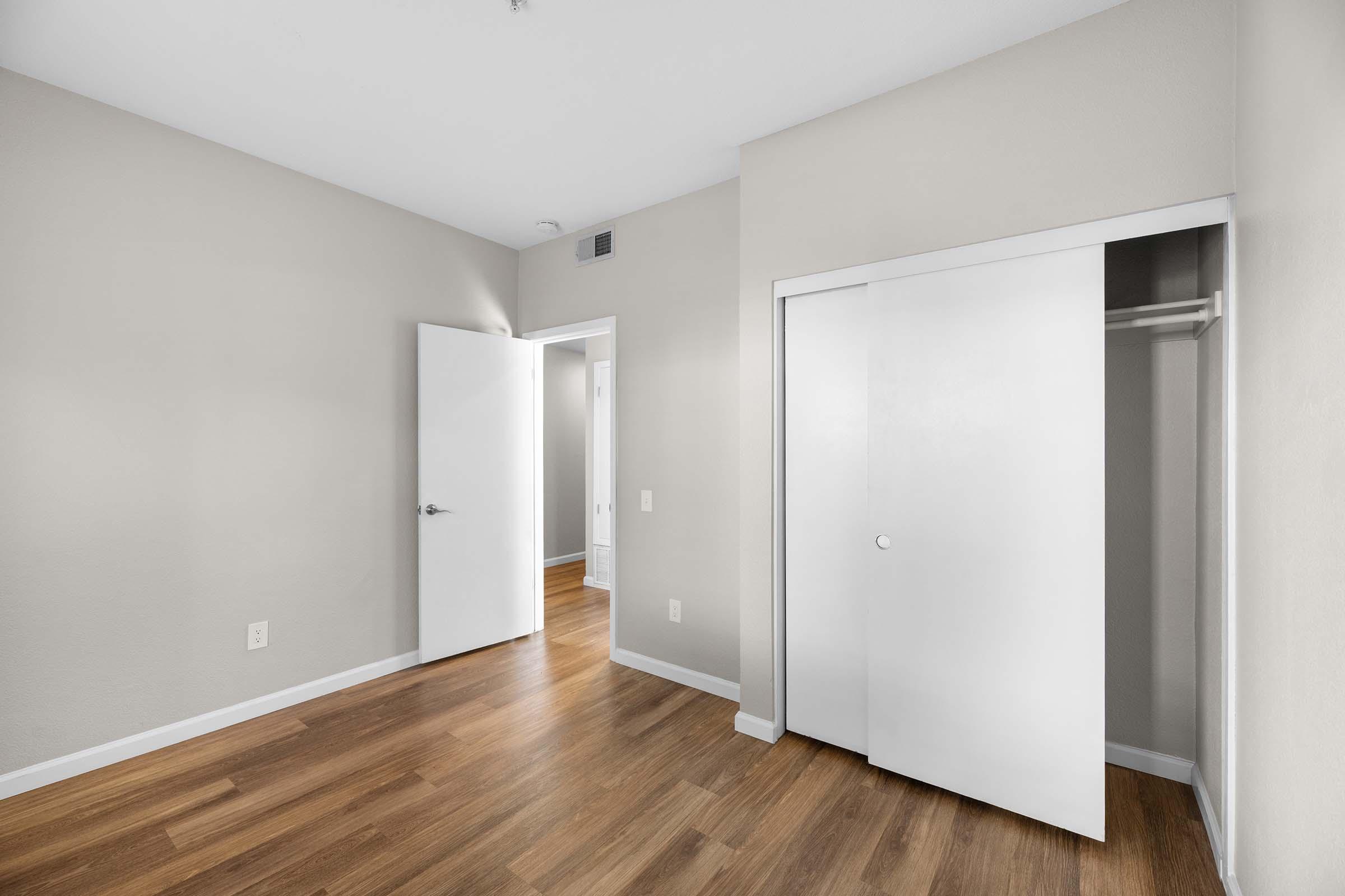 A clean, minimalistic room with light beige walls and wood flooring. It features a slightly open white door leading to another room, a closet with sliding doors, and a simple layout that emphasizes spaciousness and natural light.