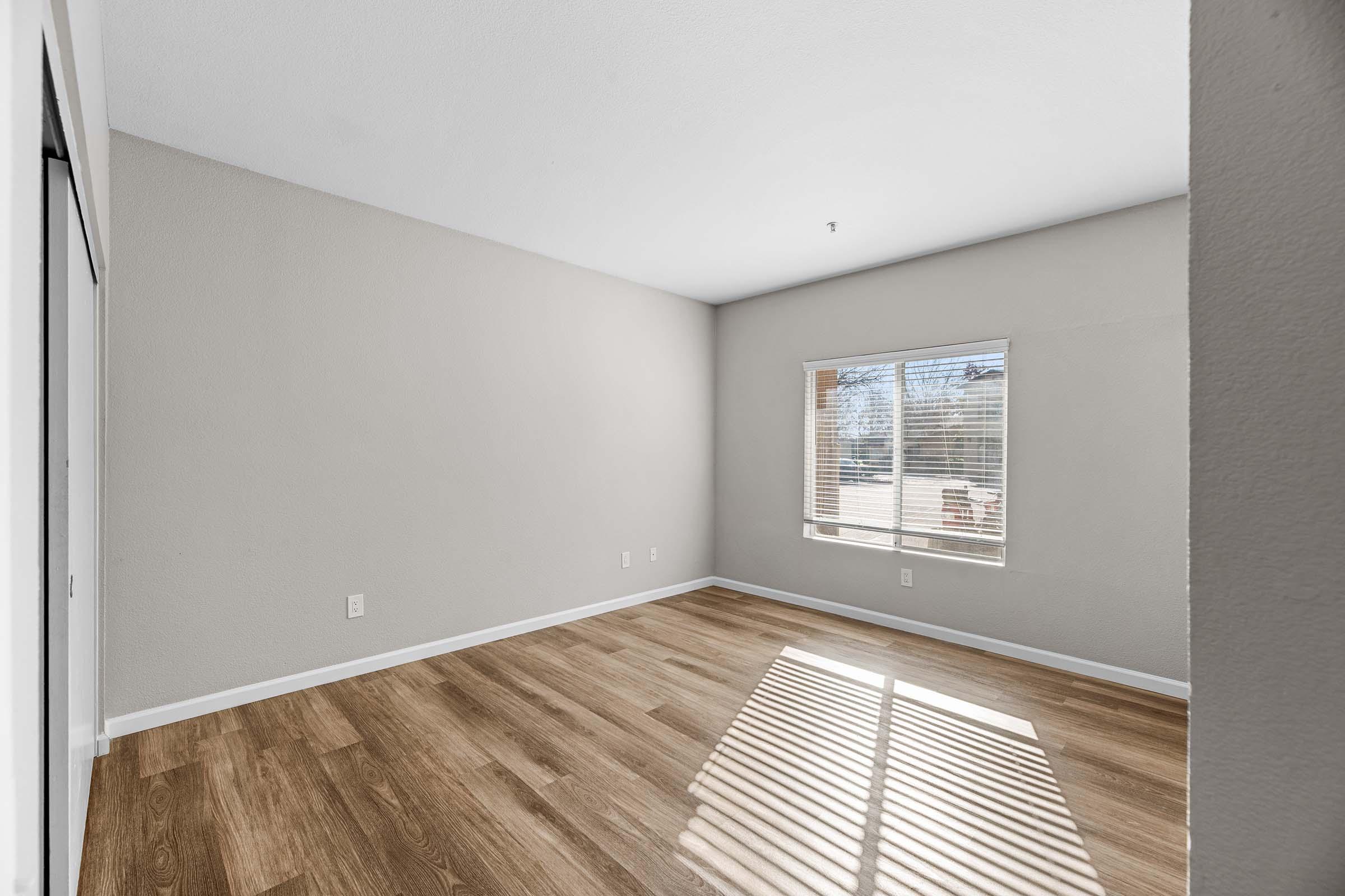 A vacant room with light-colored walls and wooden flooring. A window with blinds lets in natural light, creating shadows on the floor. The space is empty, giving a sense of openness, suitable for various uses or furnishings.