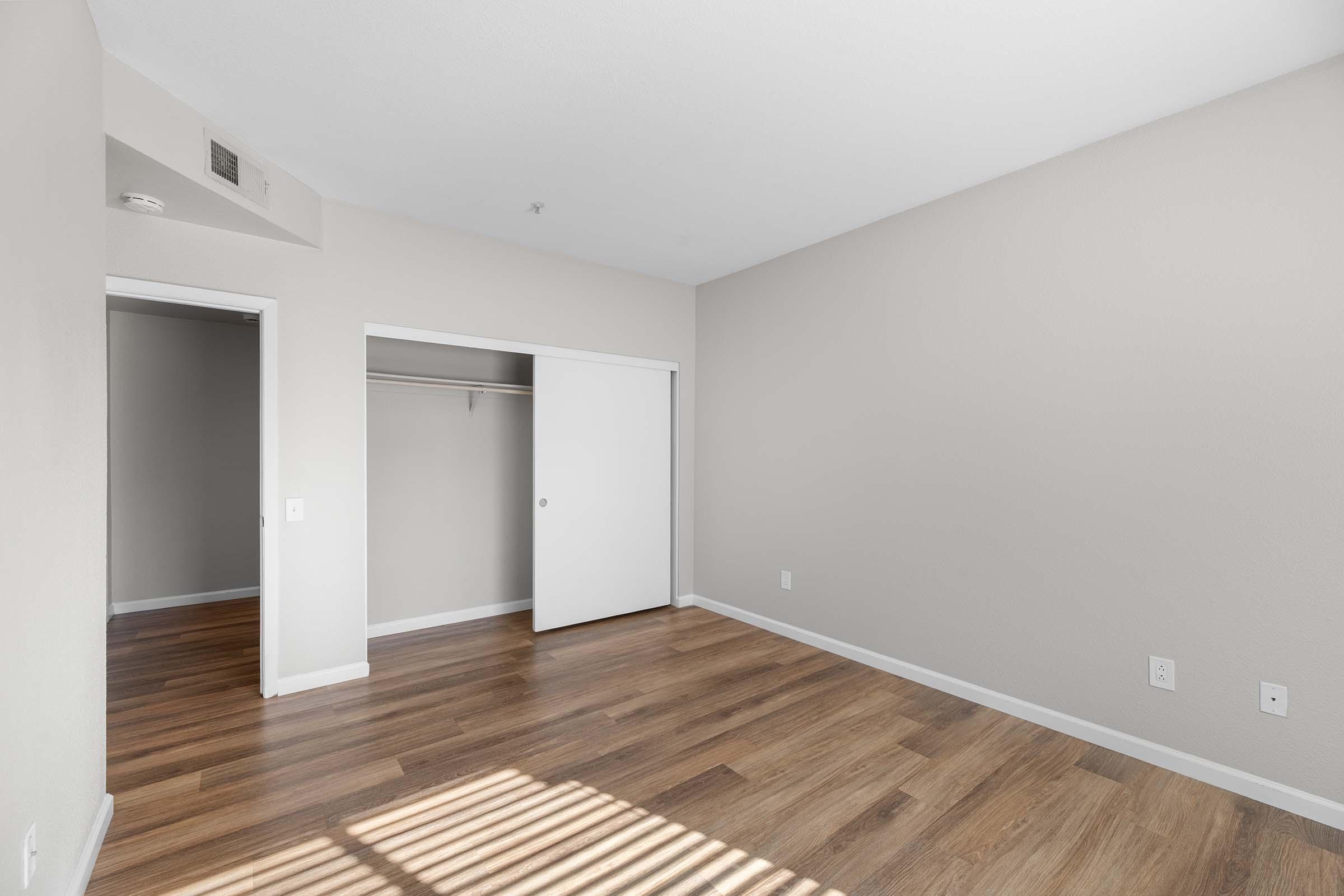 Spacious, empty room with light brown wooden flooring, light beige walls, and a white sliding closet door. The room features a large window casting shadows on the floor, adding natural light to the space. The overall atmosphere is clean and minimalistic, ideal for personalization.