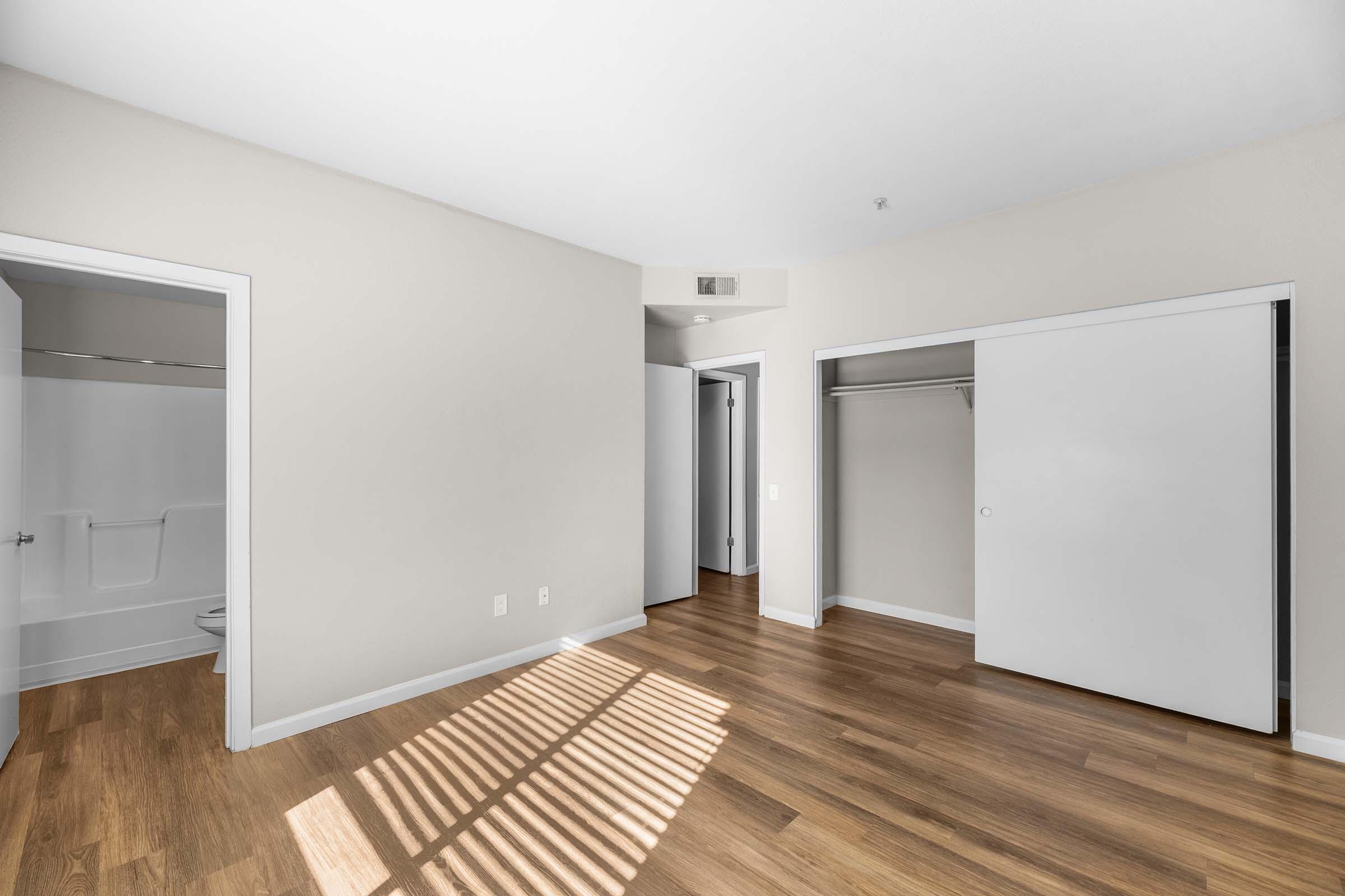 A lightly lit empty room with hardwood flooring. Two doors are visible: one leading to a bathroom and another to a closet. Sunlight casts shadows from window blinds, highlighting the walls and floor. The room features neutral-colored walls and a modern, minimalist design.