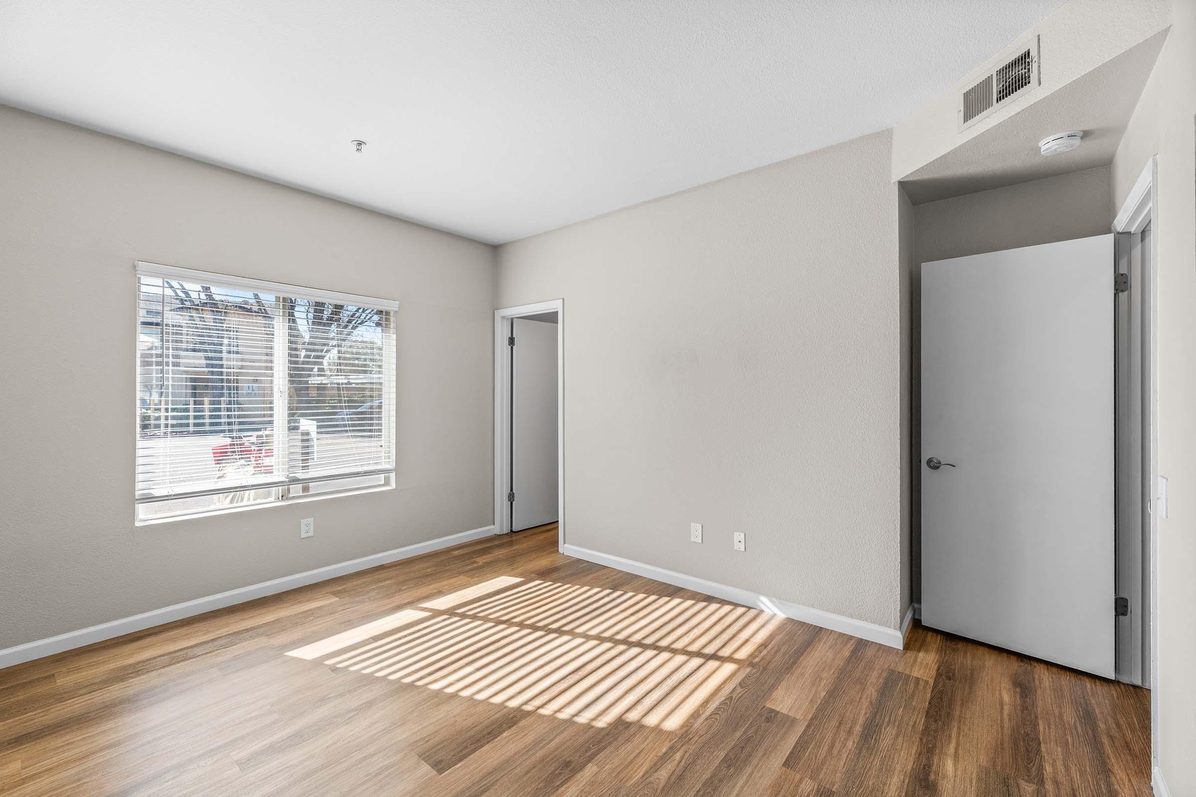 A bright, empty interior room showcasing light gray walls, a large window with blinds allowing natural light to enter, and light brown laminate flooring. There is a door leading to another space visible in the background, creating a spacious and inviting atmosphere.