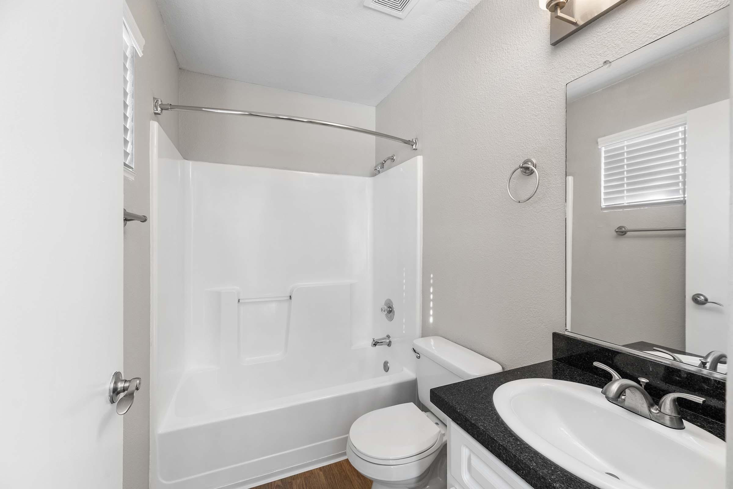 A modern bathroom featuring a white bathtub with a shower, a black countertop sink, a toilet, and a large mirror. The walls are a neutral color, and there are small windows allowing natural light. The flooring is a wood-like design, adding warmth to the space.