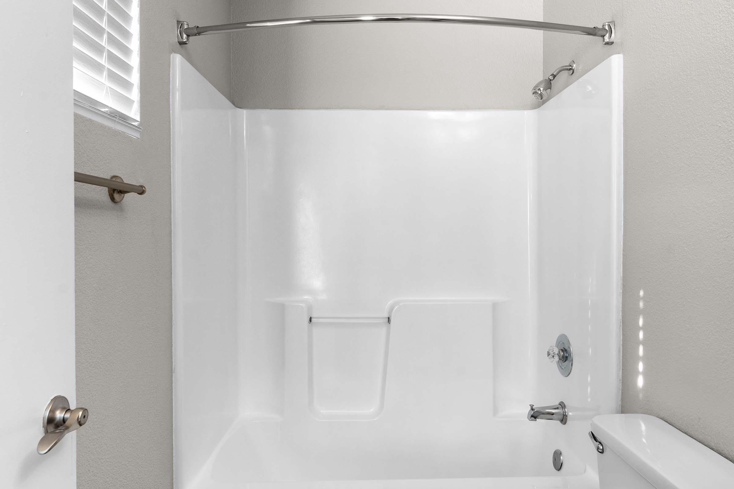 A clean and minimalist bathroom featuring a white shower enclosure with a curved shower rod, a tub, and a small window allowing natural light. The walls are a light gray, enhancing the bright and spacious feel of the area.