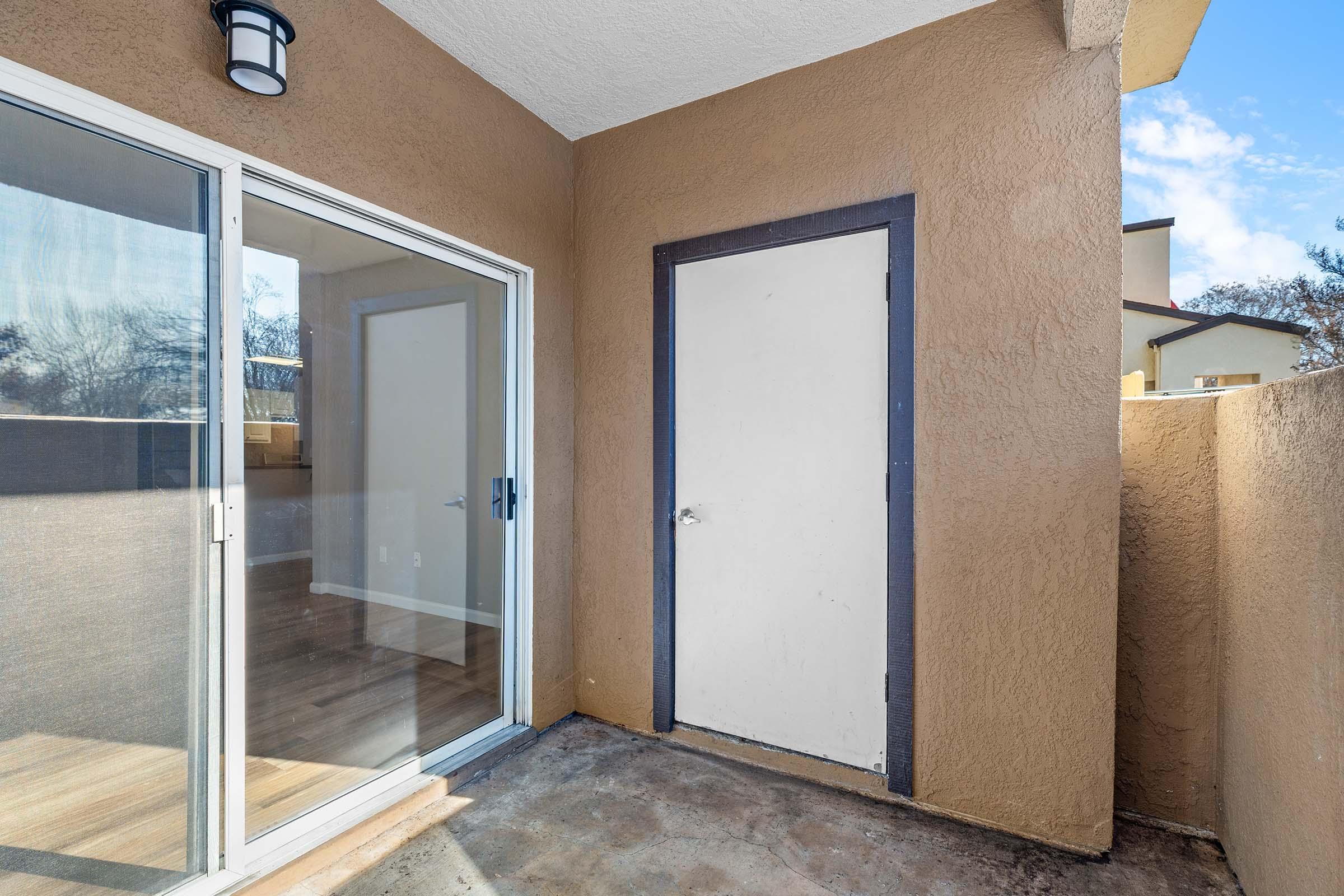 A small patio area with a sliding glass door leading indoors and a plain white door on the adjacent wall. The walls are painted a warm beige, and the floor appears to be concrete. Natural light filters in, highlighting the space's simplicity and potential for outdoor use.