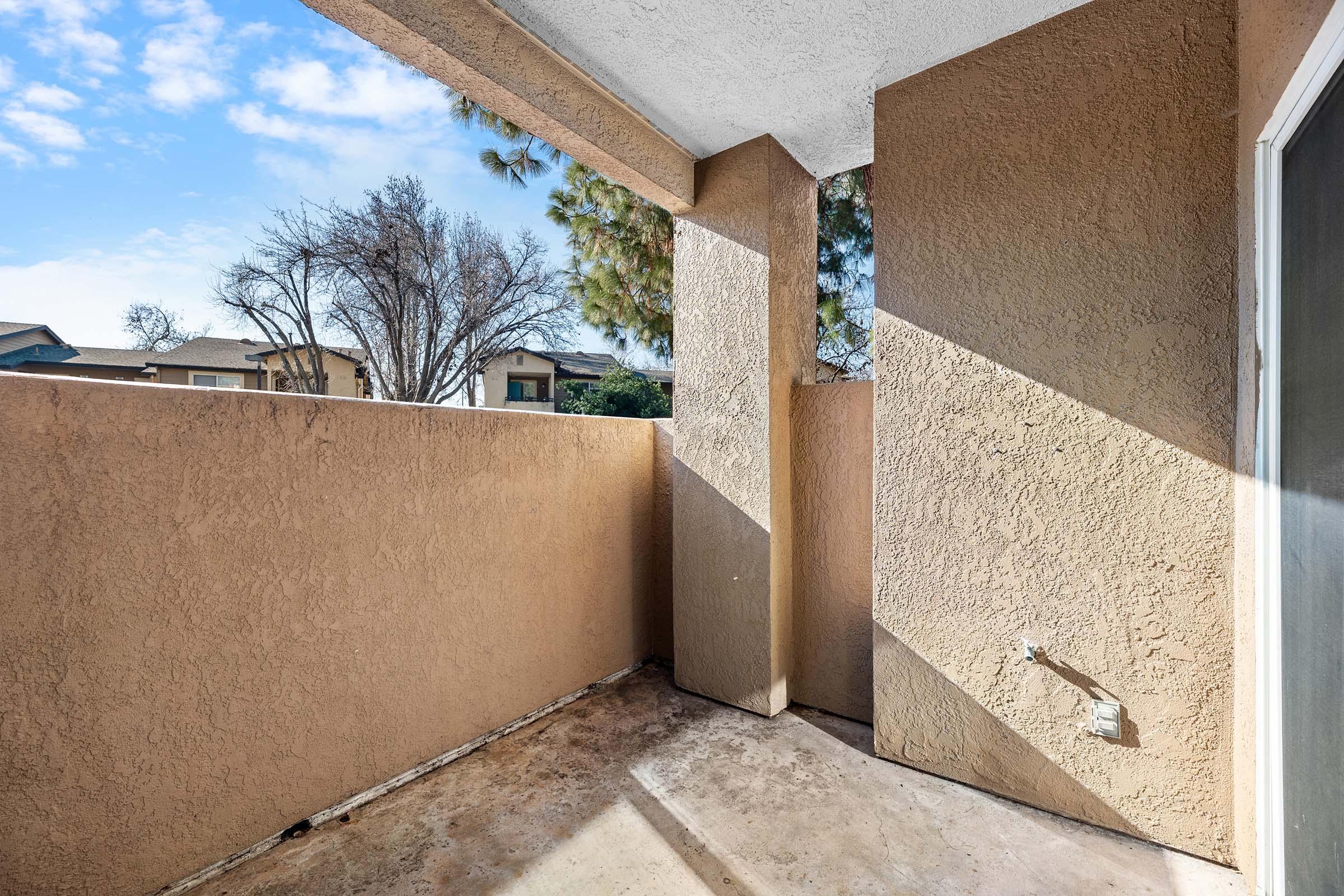 View of a small balcony with beige walls, featuring a textured surface. The corner has a vertical pillar, and sunlight is casting diagonal shadows across the floor. In the background, there are sparse trees and a glimpse of neighboring buildings under a clear blue sky.