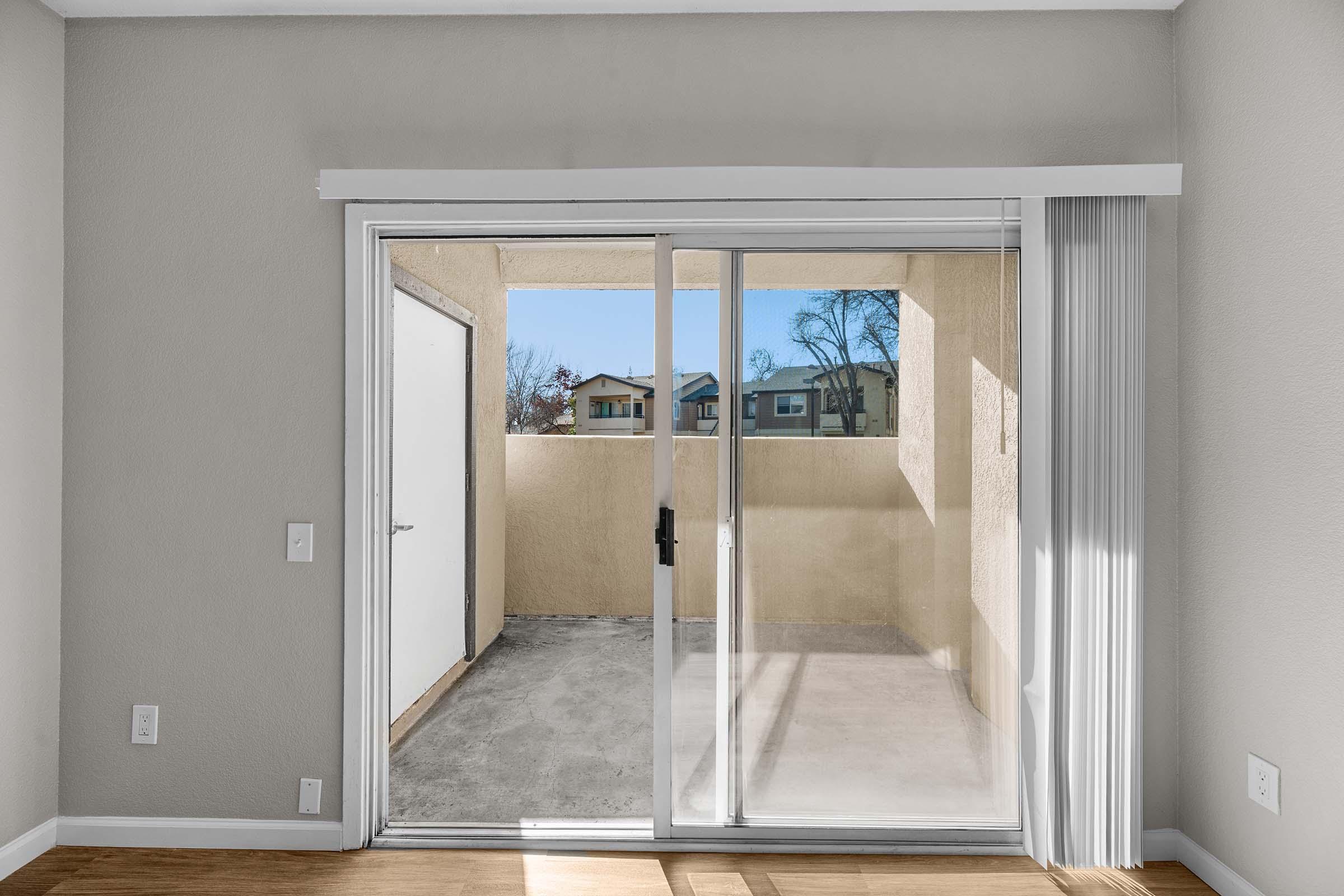Interior view of a living space featuring a sliding glass door leading to a small outdoor patio area, with light-colored walls and a wooden floor. Sunlight streams in, illuminating the room. The patio is enclosed by beige walls, and there are views of neighboring buildings from the door.