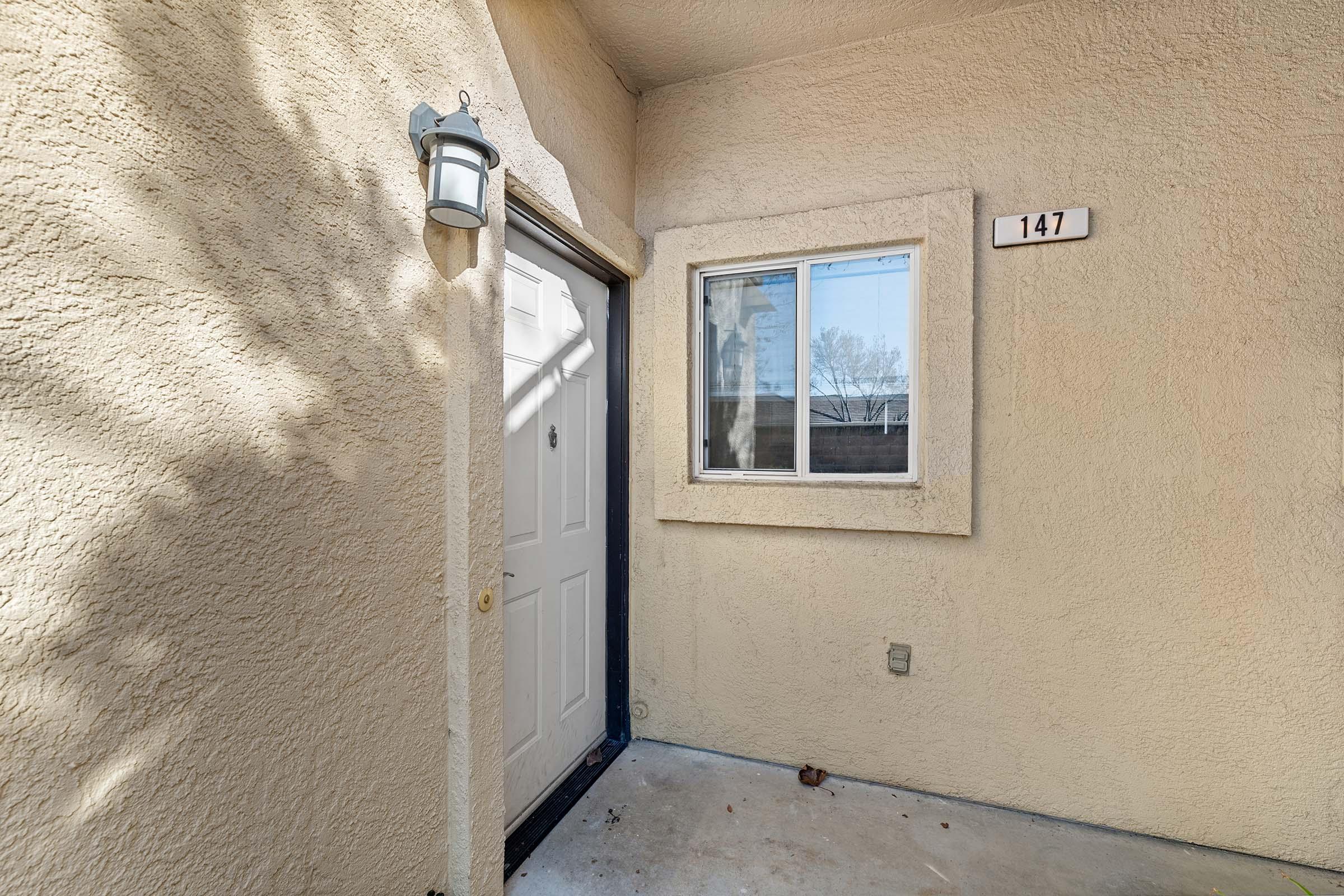 Exterior view of a light-colored wall with a closed white door and a window beside it. The door is marked with the number 147. A light fixture is mounted above the door, and a tree's shadow is partially visible on the wall. The area appears well-maintained and inviting.