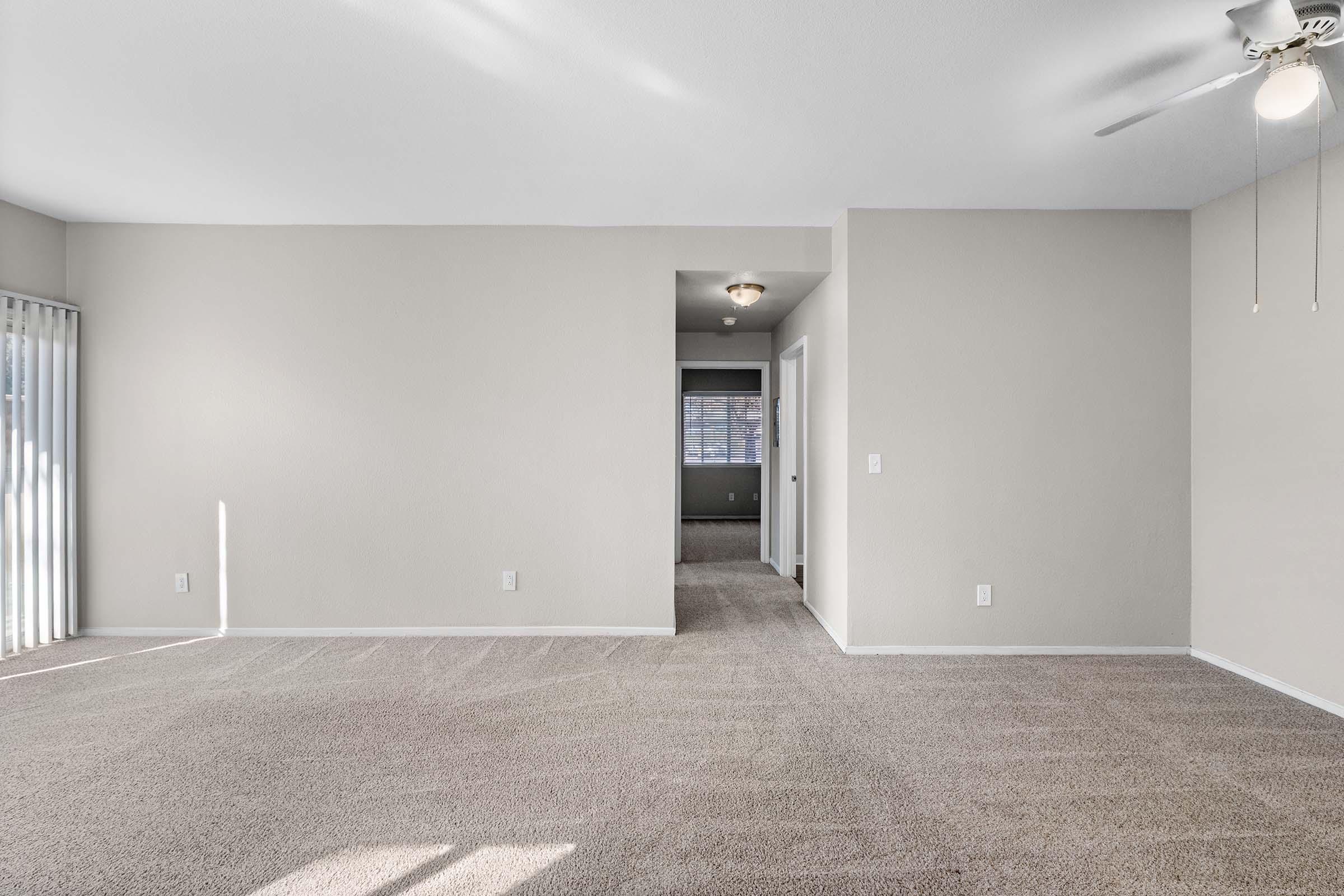 Empty living space with beige walls and carpeted flooring, featuring a ceiling fan. A doorway leads to another room, while sliding glass doors are visible at one side, allowing natural light to enter. The overall ambiance is bright and spacious.