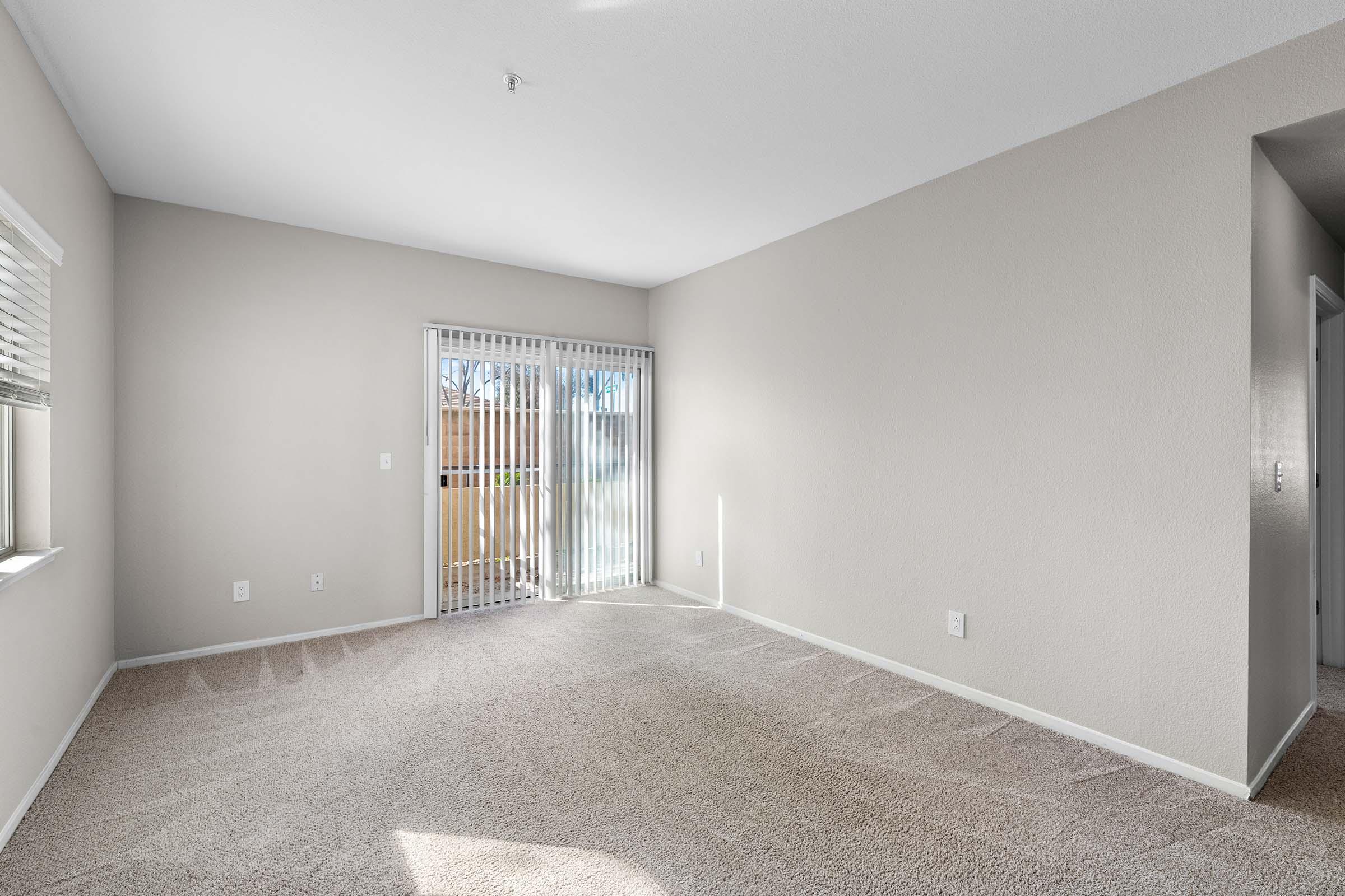 Empty room with light beige walls and a carpeted floor. A large sliding glass door leads to an outdoor space, allowing natural light to enter. The room has minimal furnishings and decor, emphasizing a clean and open layout.