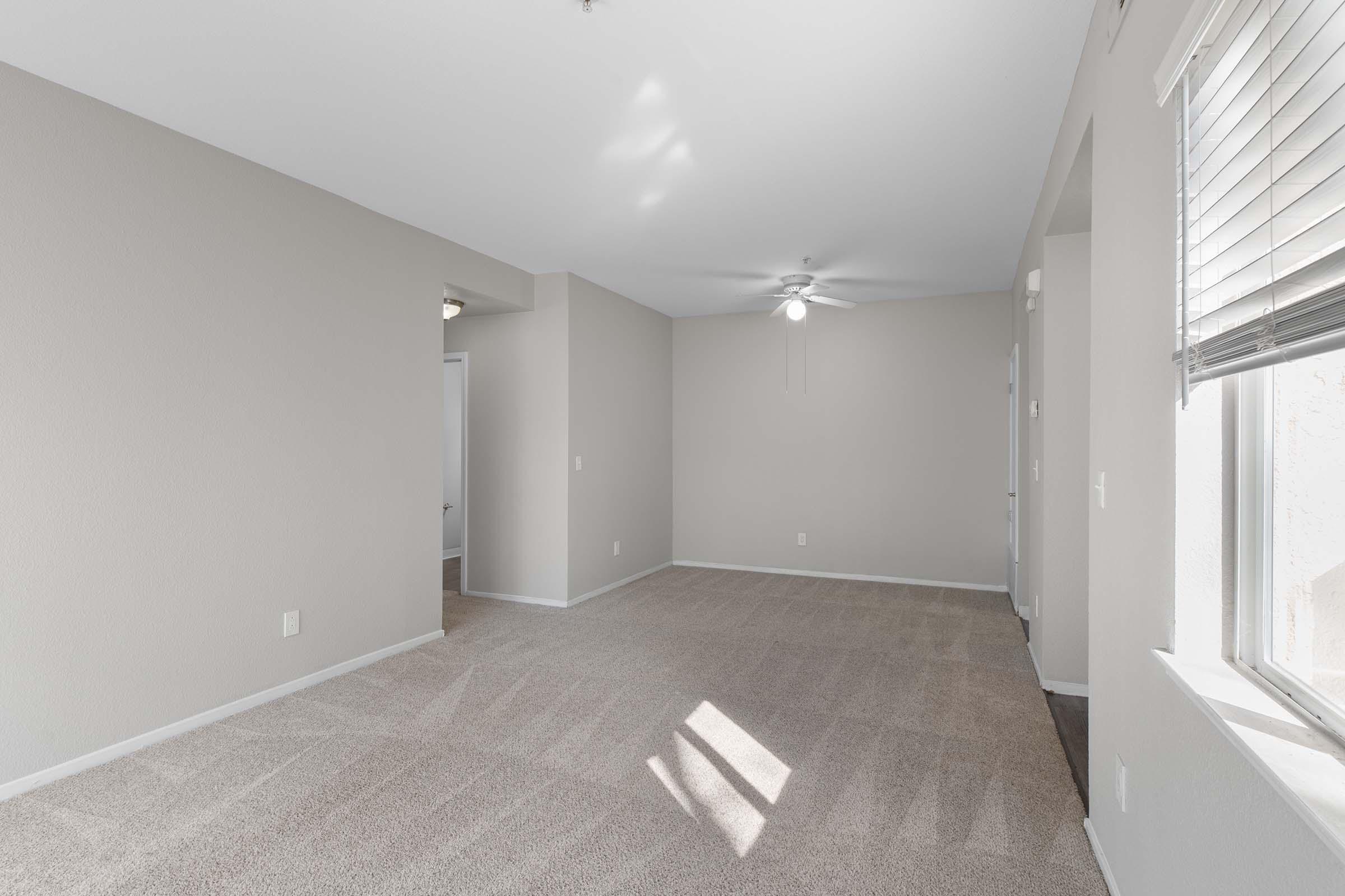 A spacious, empty living room with light beige walls and a light carpet. Large windows allow natural light to fill the room, creating a bright and inviting atmosphere. A ceiling fan is visible, and doorways lead to other areas of the home.