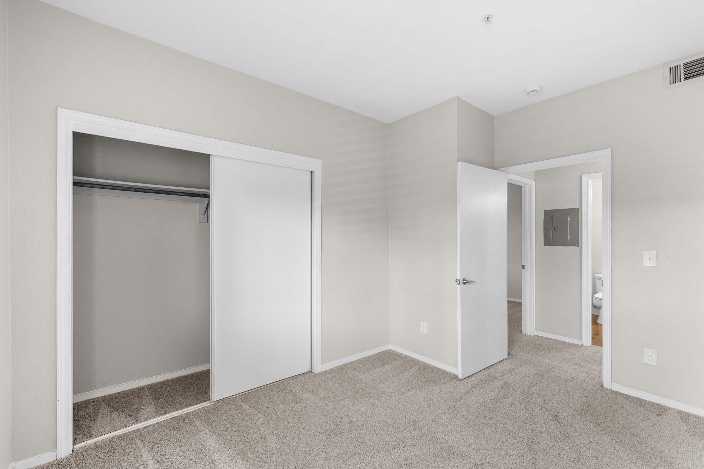 An empty room with light beige walls and carpet. There are two closed doors, one leading to a small closet and the other likely to a hallway or additional room. A bathroom can be seen through an open door in the background. The overall space is well-lit and neat.