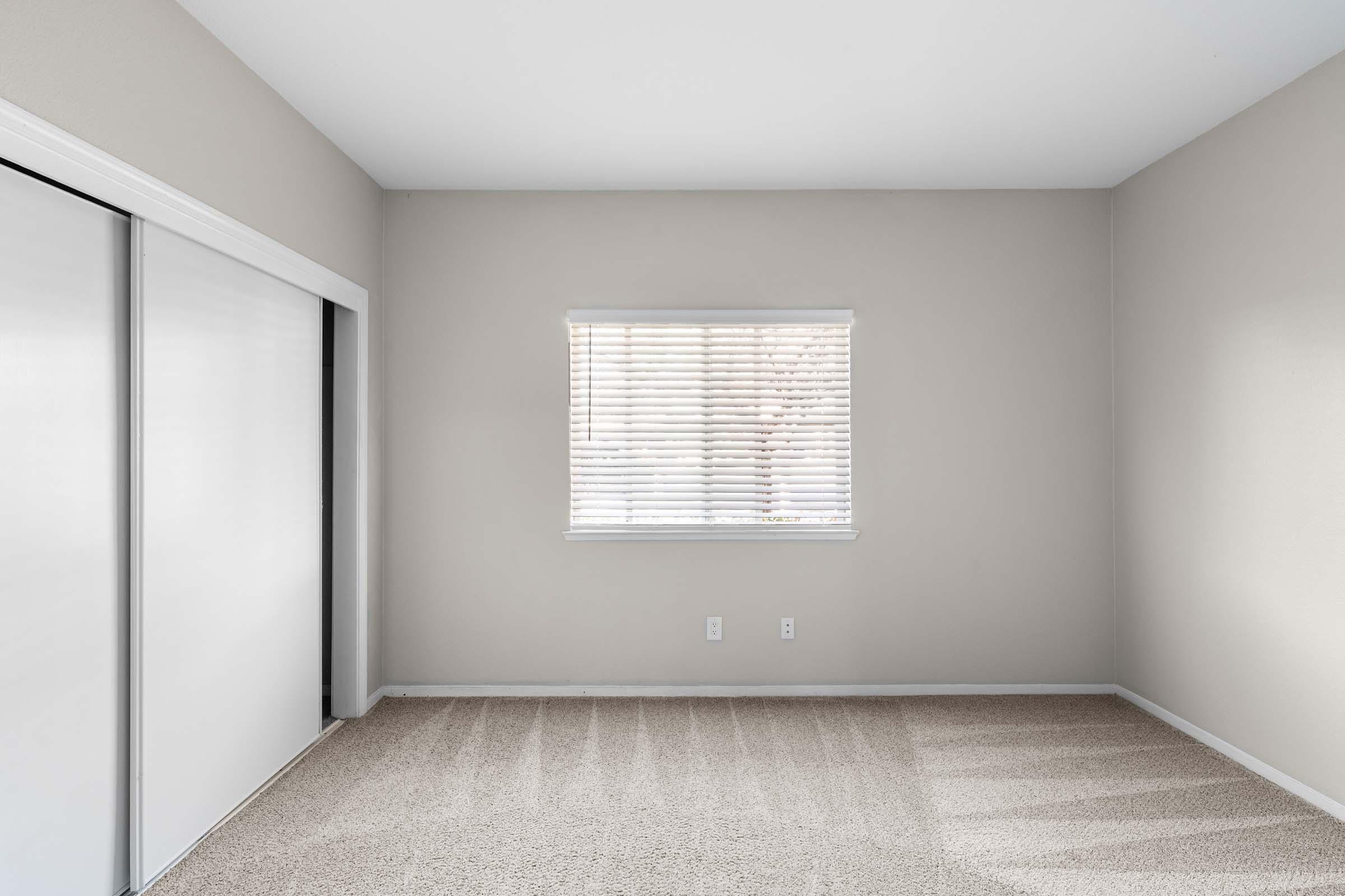 A well-lit, empty room with beige walls and beige carpet. There is a window with blinds on one wall and two sliding closet doors on the opposite wall. The space appears clean and ready for furnishings.