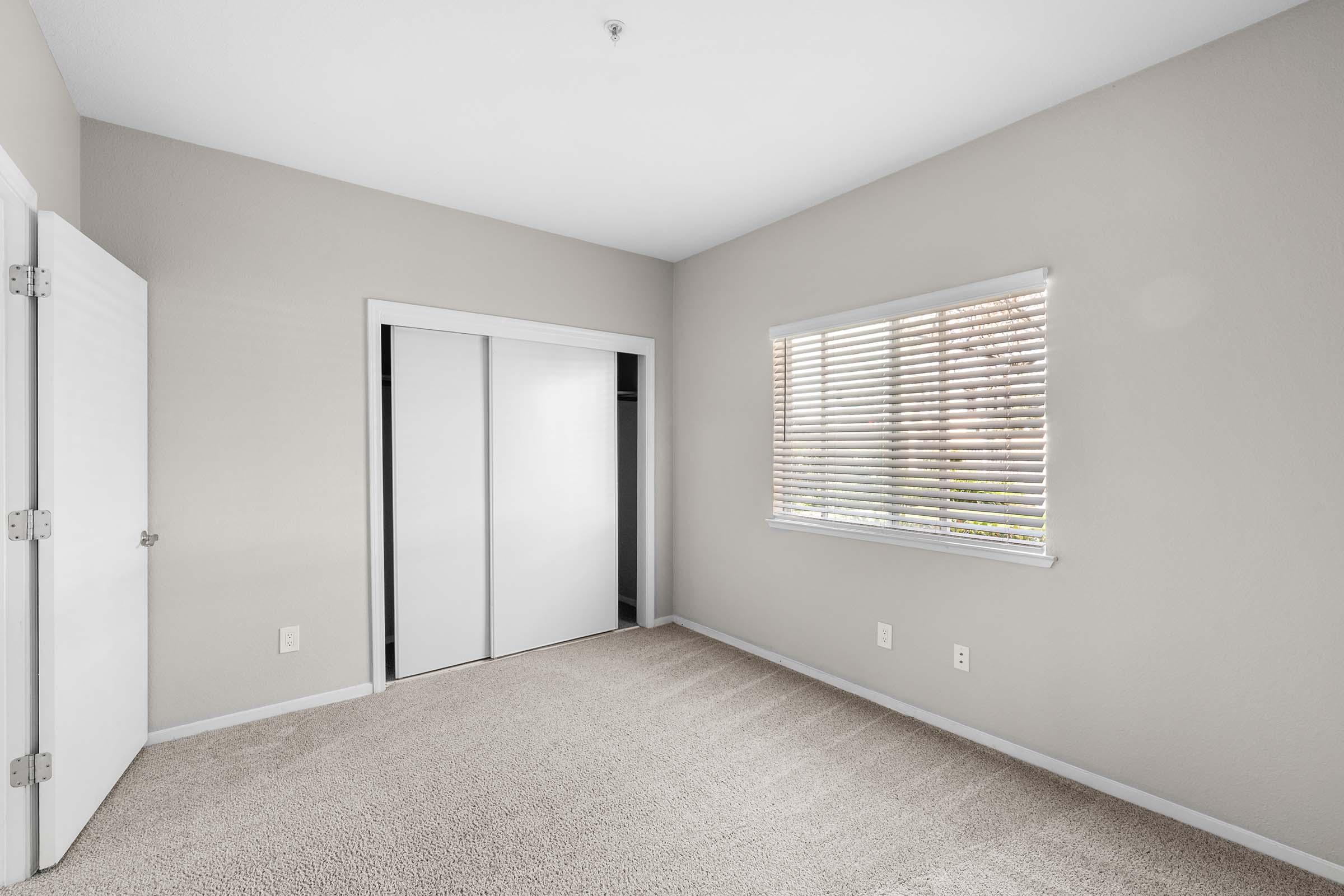 A vacant room featuring beige walls and carpet, with a window covered by blinds allowing natural light to enter. There are white double doors leading to a closet on one side, and the overall space appears clean and uncluttered, suitable for various uses.