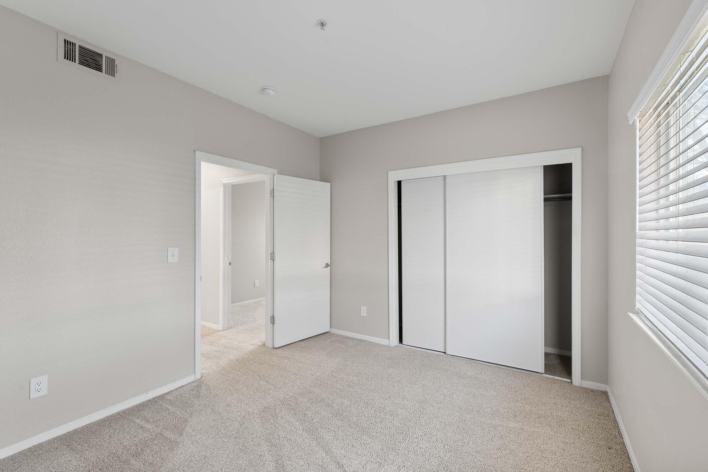 A vacant bedroom featuring light-colored walls, carpeted floors, and a closet with sliding doors. There is a doorway leading to another room, and a window providing natural light. The space is clean and minimalistic, suitable for personalization.