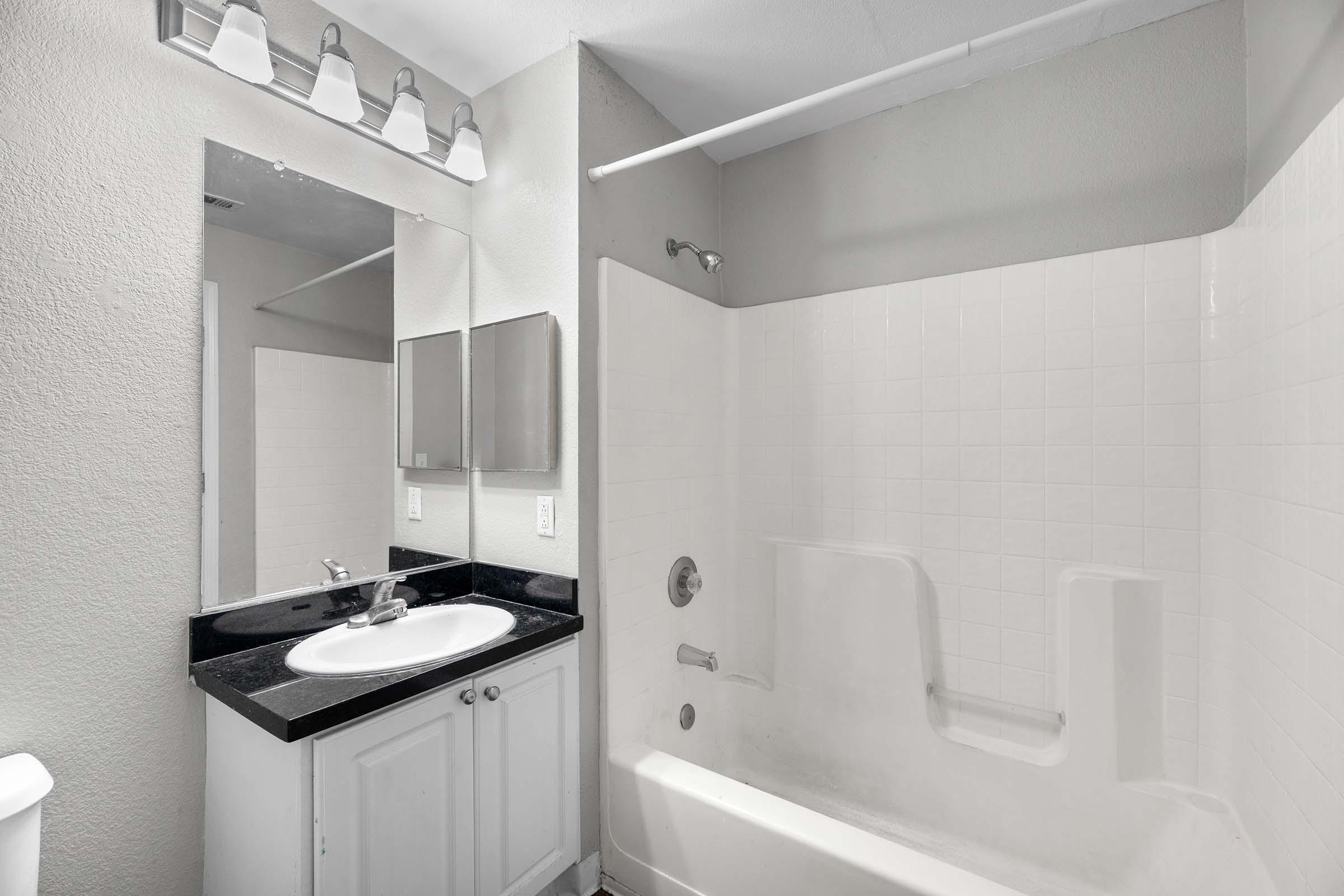 A clean and modern bathroom featuring a white bathtub with a built-in shower, a dark countertop with a sink, two mirrors, and bright lighting. The walls are painted a light gray, creating a spacious and inviting atmosphere. A toilet is visible in the corner, completing the functional design.