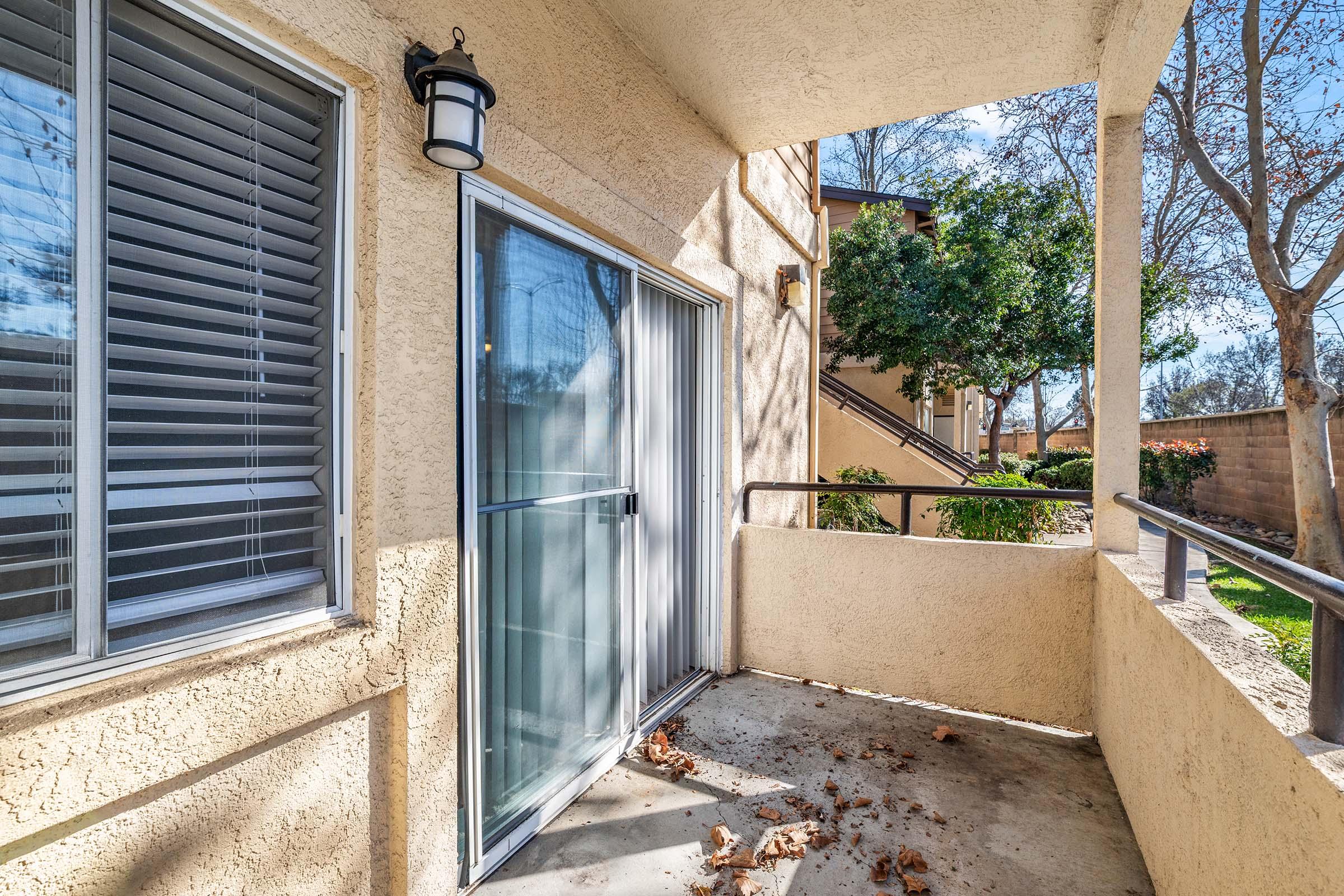 A view of a small balcony with a sliding glass door, surrounded by beige walls. The floor is partially covered with dry leaves, and there are trees and plants visible in the background. The balcony features a light fixture and overlooks a landscaped area.