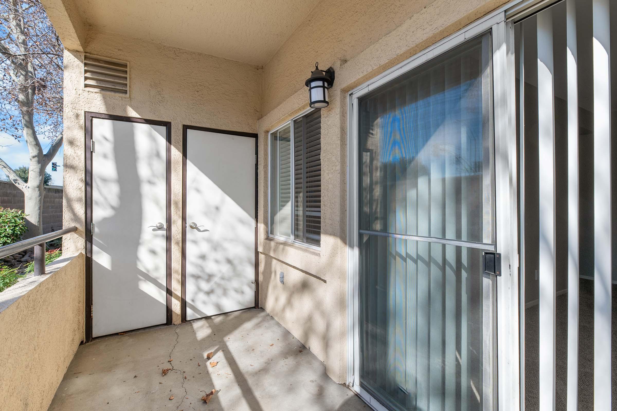 A small outdoor balcony area with a concrete floor, featuring two doors—one white and another with glass panels. Sunlight casts shadows on the walls, and there are plants visible in the background, creating a cozy atmosphere.