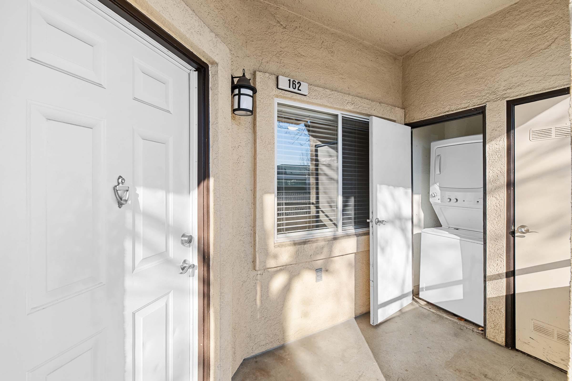 An outdoor view of a small apartment entrance featuring two white doors, one labeled "162." A window nearby lets in light, and a stackable washer and dryer is visible inside the unit. The setting has a neutral color scheme and is well-lit, creating a welcoming atmosphere.