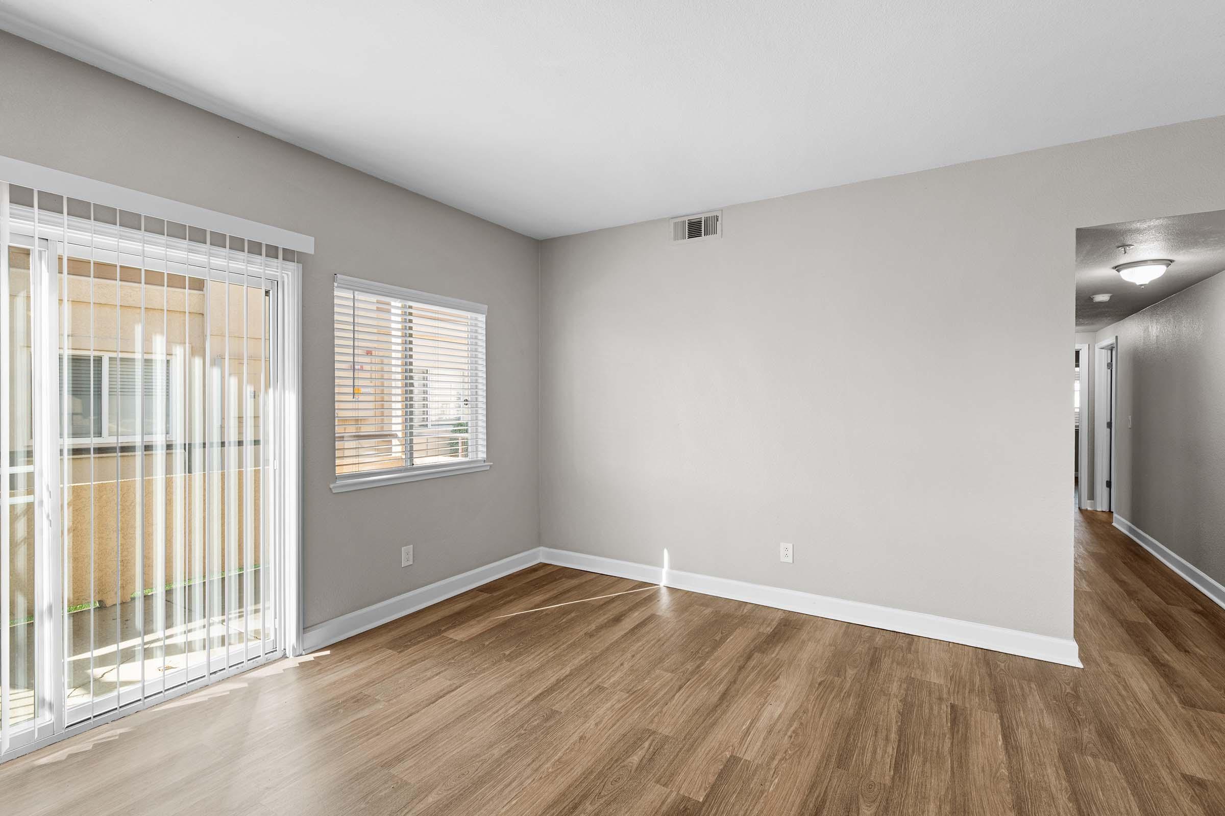 Interior view of a spacious, empty room with light beige walls and hardwood flooring. A large window with blinds allows natural light to enter from outside, revealing a patio area. A doorway on the right side leads to another part of the home, while the room appears freshly painted and well-lit.