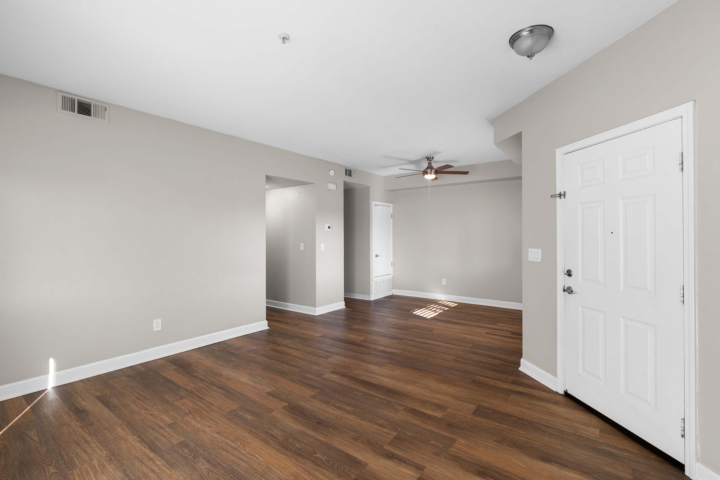 A spacious living area with light-colored walls and dark hardwood flooring. The room features a ceiling fan and a front door with a white frame. To the side, there's an entrance to another room or hallway. Natural light streams in, highlighting the open space and neutral decor, creating a welcoming atmosphere.