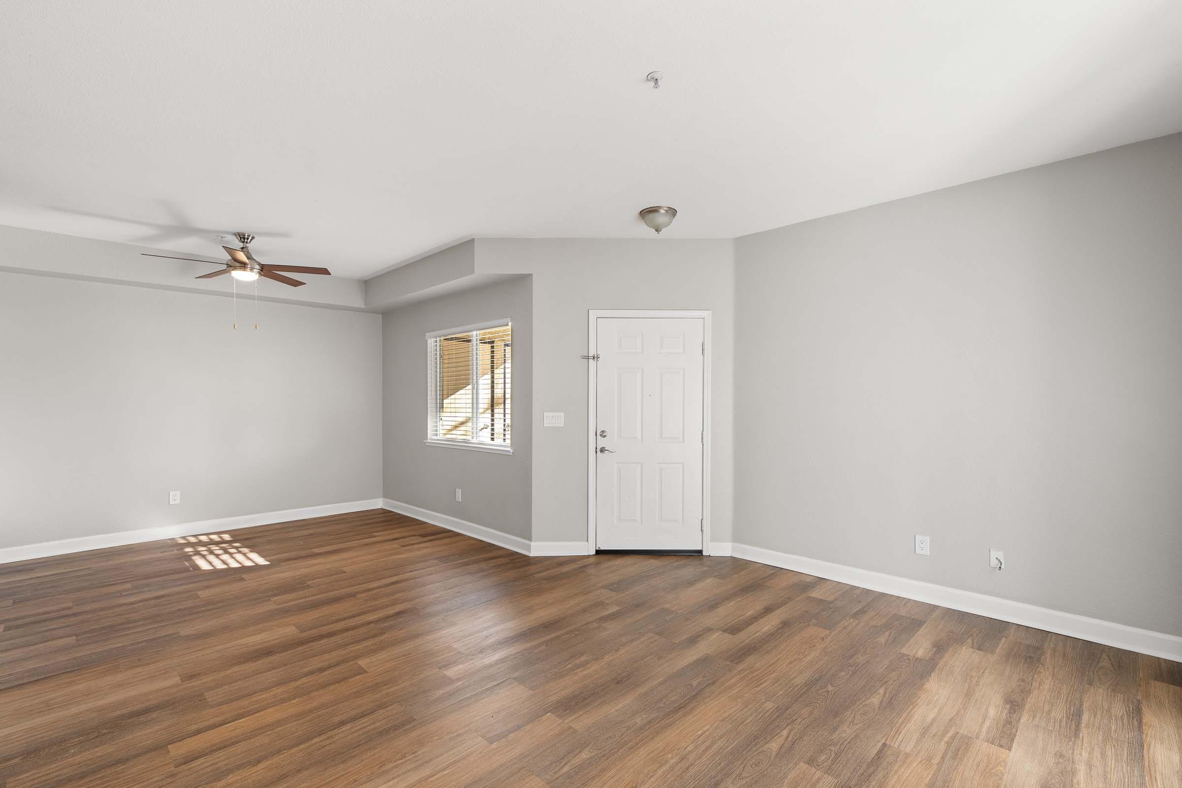 A spacious, empty room with light gray walls and a ceiling fan. The floor is made of light wood. There is a closed door with a white frame on one side and a window letting in natural light.