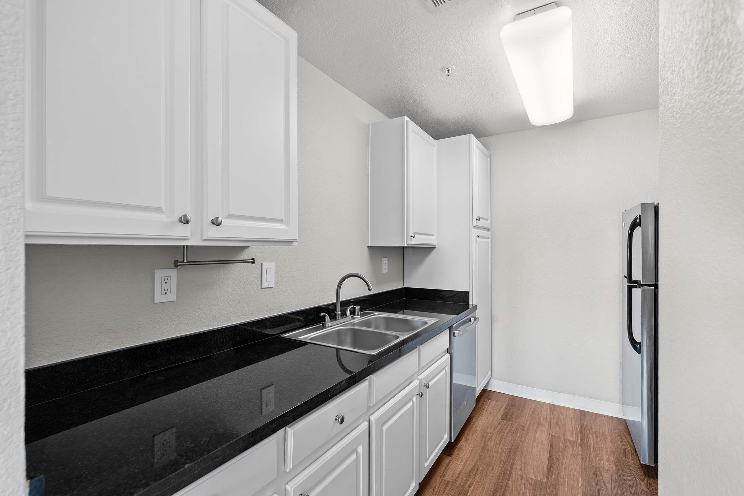 A modern kitchen featuring white cabinets, a black countertop, stainless steel sink, and refrigerator. The space has an open layout with wood flooring and soft lighting from a ceiling fixture. The walls are painted light colors, creating a bright and inviting atmosphere.