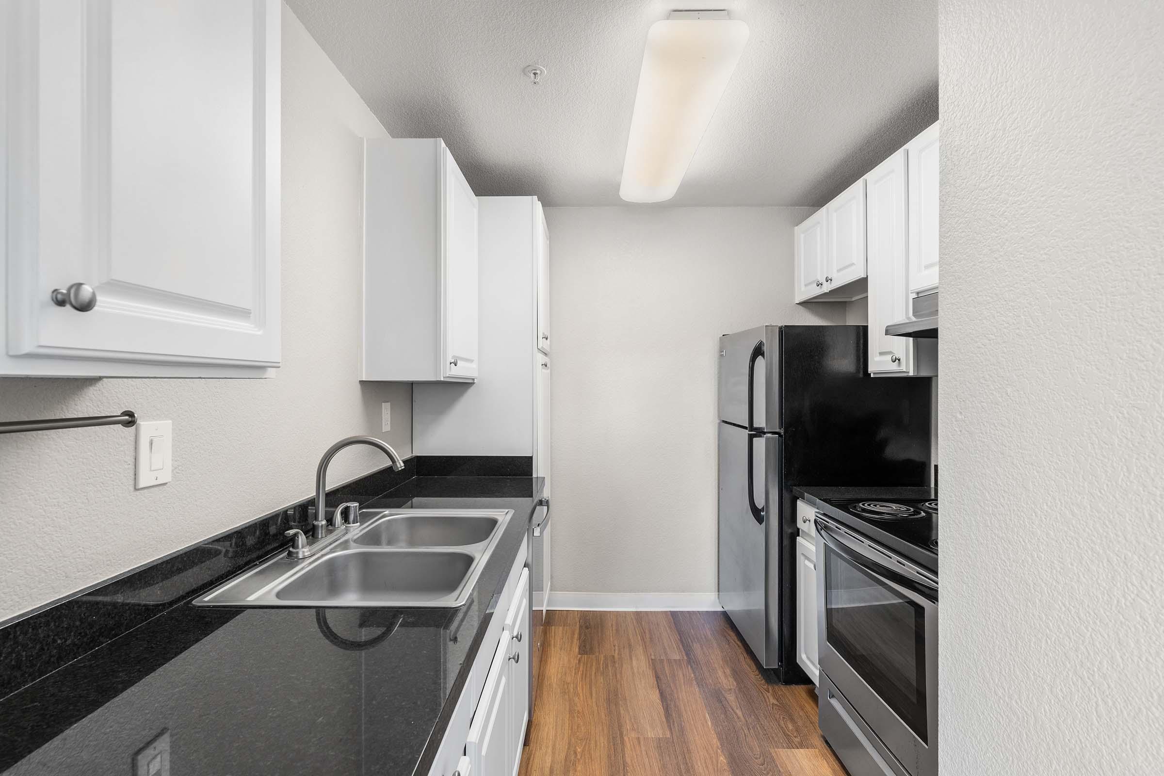 Modern kitchen featuring white cabinets, a black countertop, stainless steel sink, and a mix of black and stainless steel appliances, including a refrigerator, oven, and stove. The space is well-lit with a ceiling light and has wooden flooring, creating a clean and inviting atmosphere.