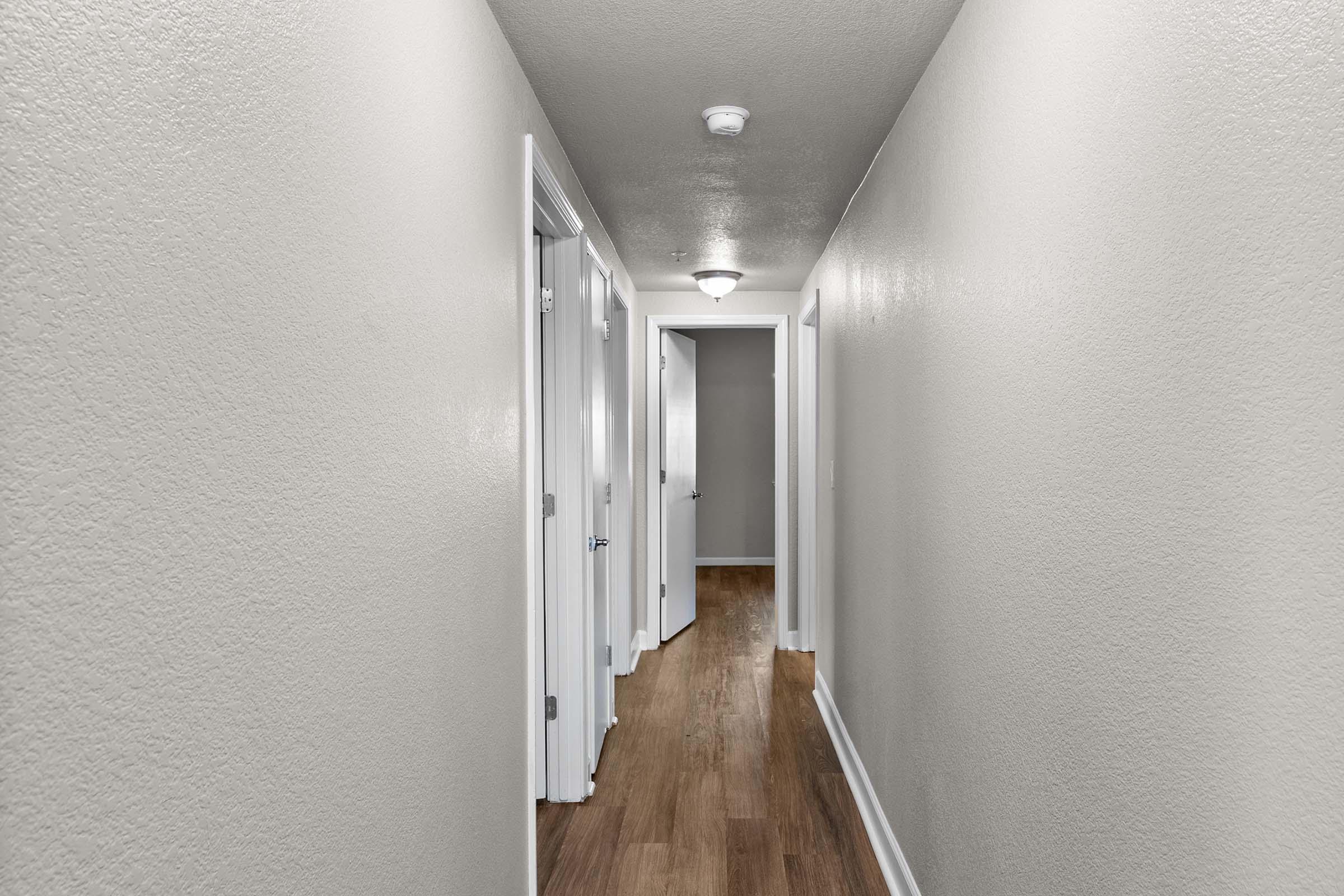 A narrow hallway with light gray walls and wooden flooring. Several doors are visible on both sides, leading to different rooms. The ceiling is equipped with a round light fixture, adding to the bright and spacious feel of the corridor.