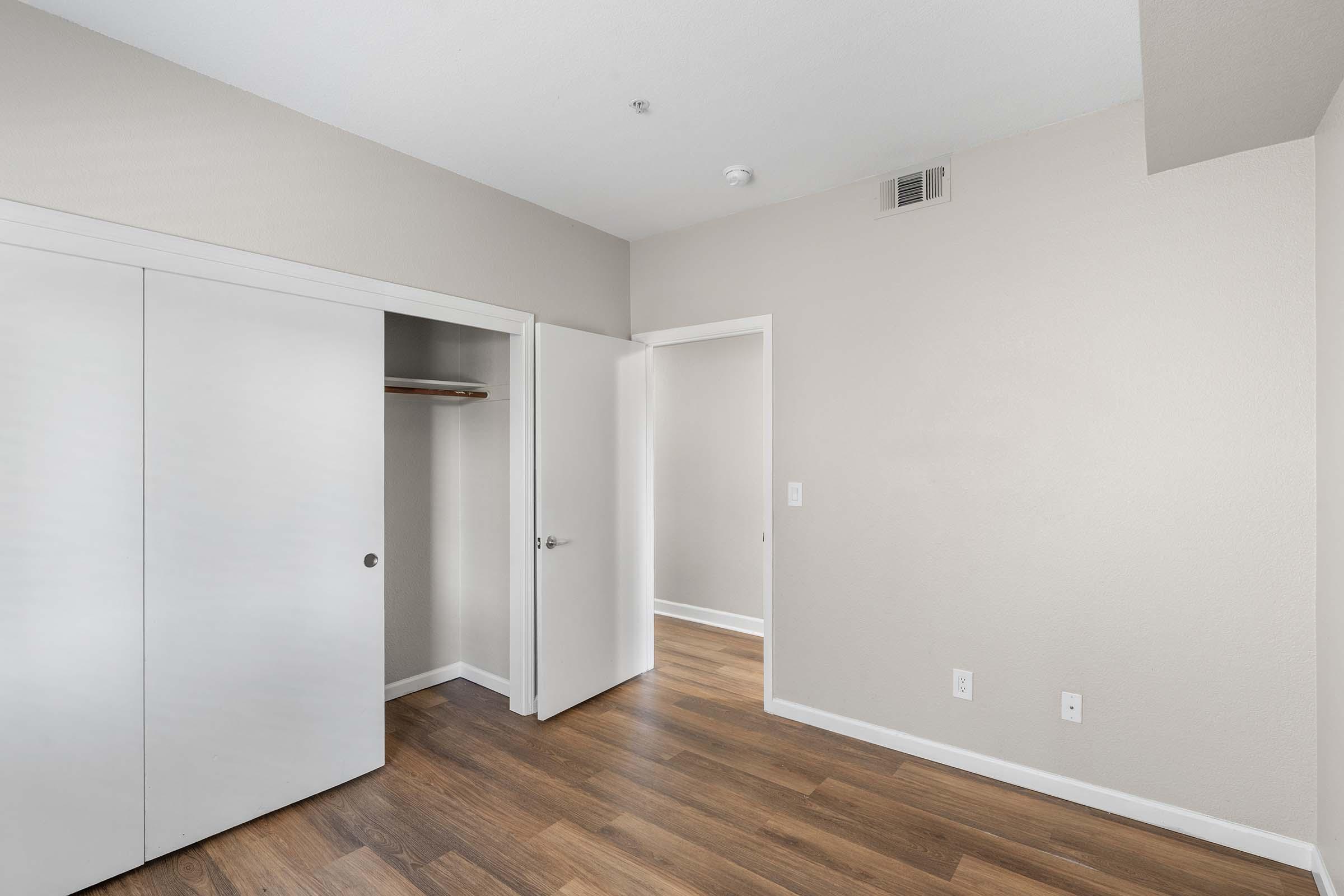 Empty room with light-colored walls and wooden flooring. There are white double sliding closet doors to the left and an open door leading to another space. Natural light fills the area, creating a bright and inviting atmosphere.