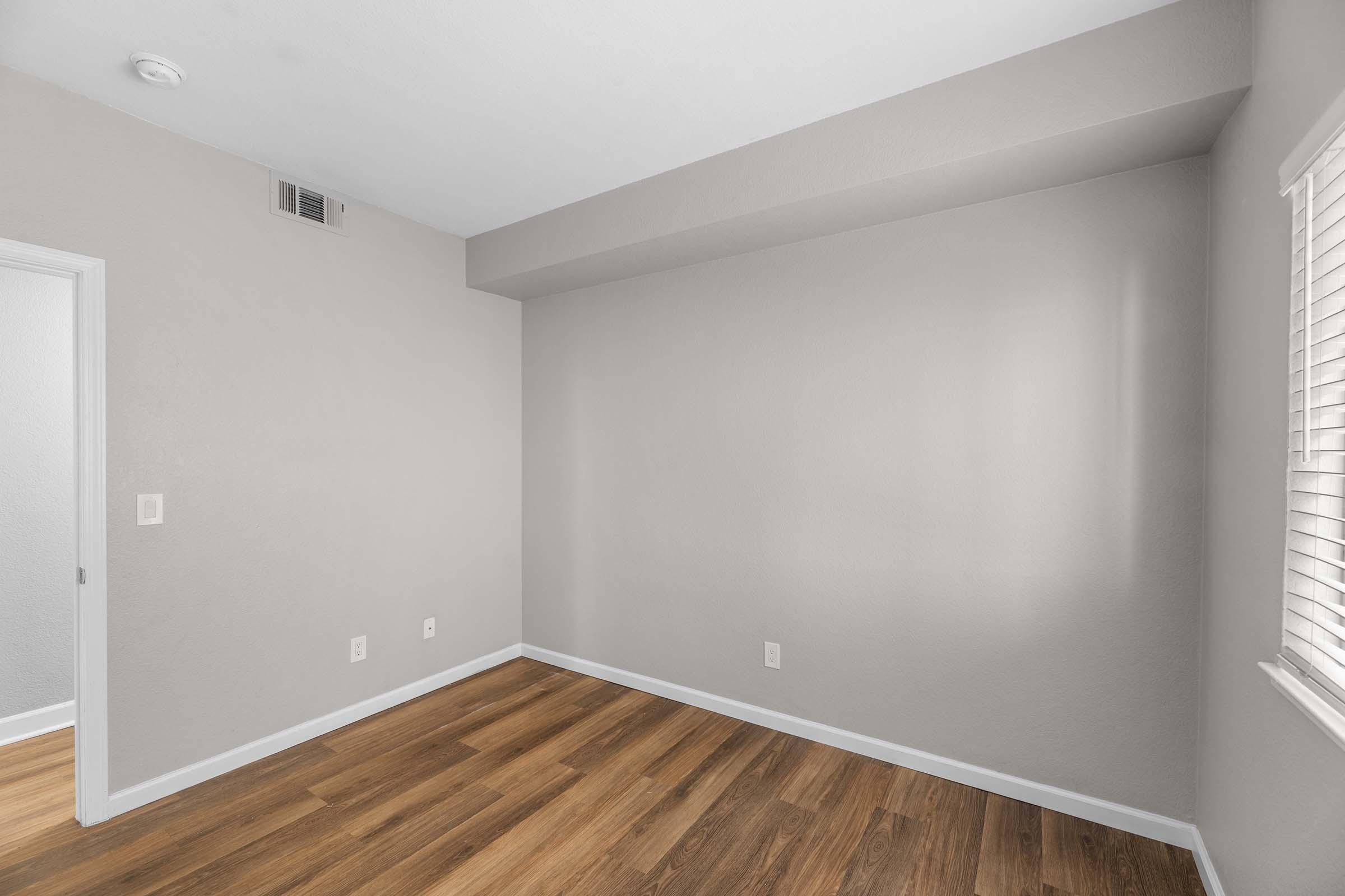 Empty room with light gray walls and wooden flooring. One wall has a window with white blinds, and there is a doorway leading to another space. The room is well-lit with natural light from the window, creating a spacious and airy atmosphere.