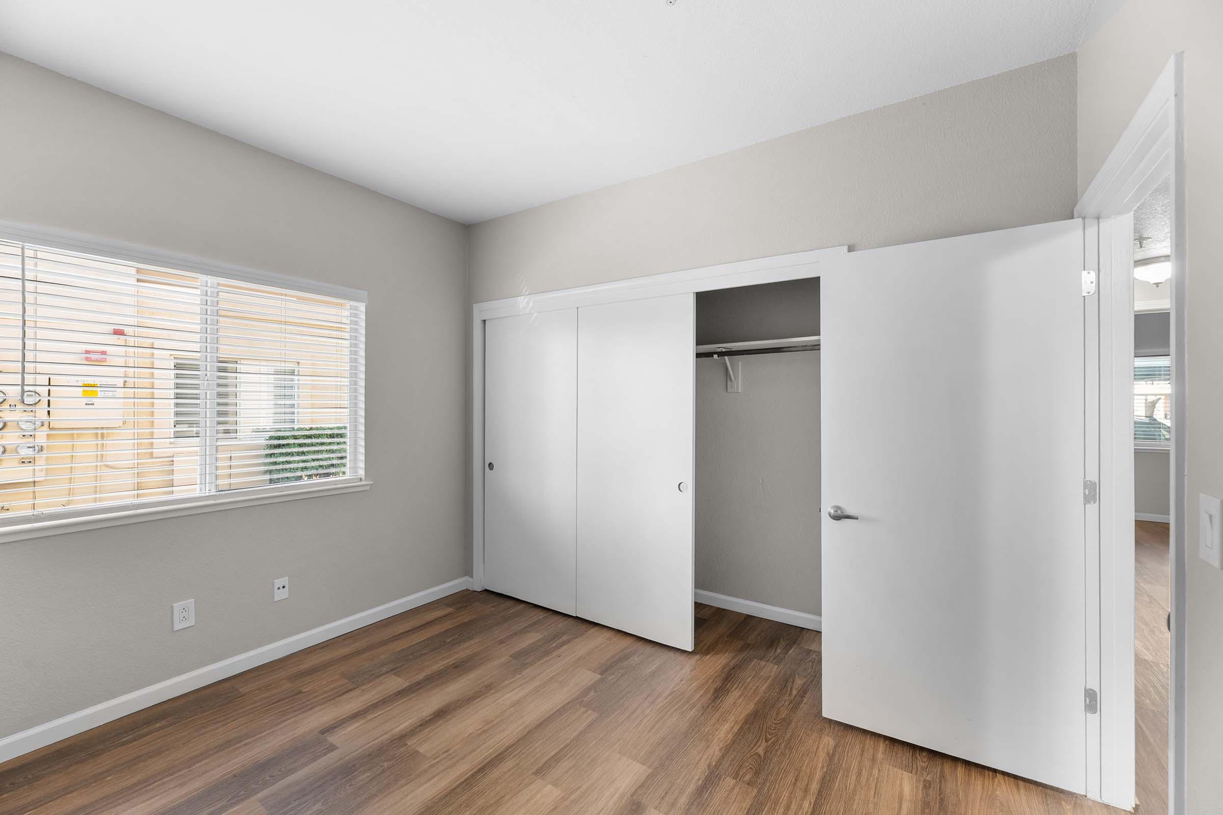 A bright, minimalist bedroom featuring light gray walls, a large window with blinds, and wooden flooring. There are two white closet doors partially open, revealing an empty closet space. The overall ambiance is clean and modern.