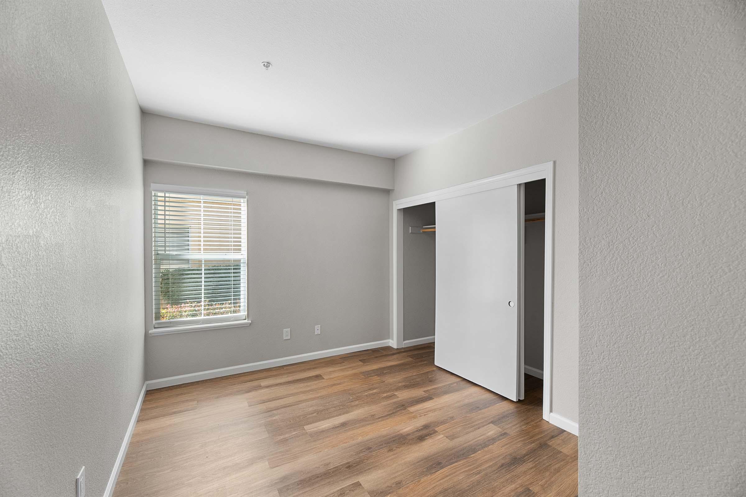A well-lit empty room with light gray walls and a large window. The flooring is a warm wood-like material. To the right, there's a closet with sliding white doors. The overall space appears clean and ready for furnishings.