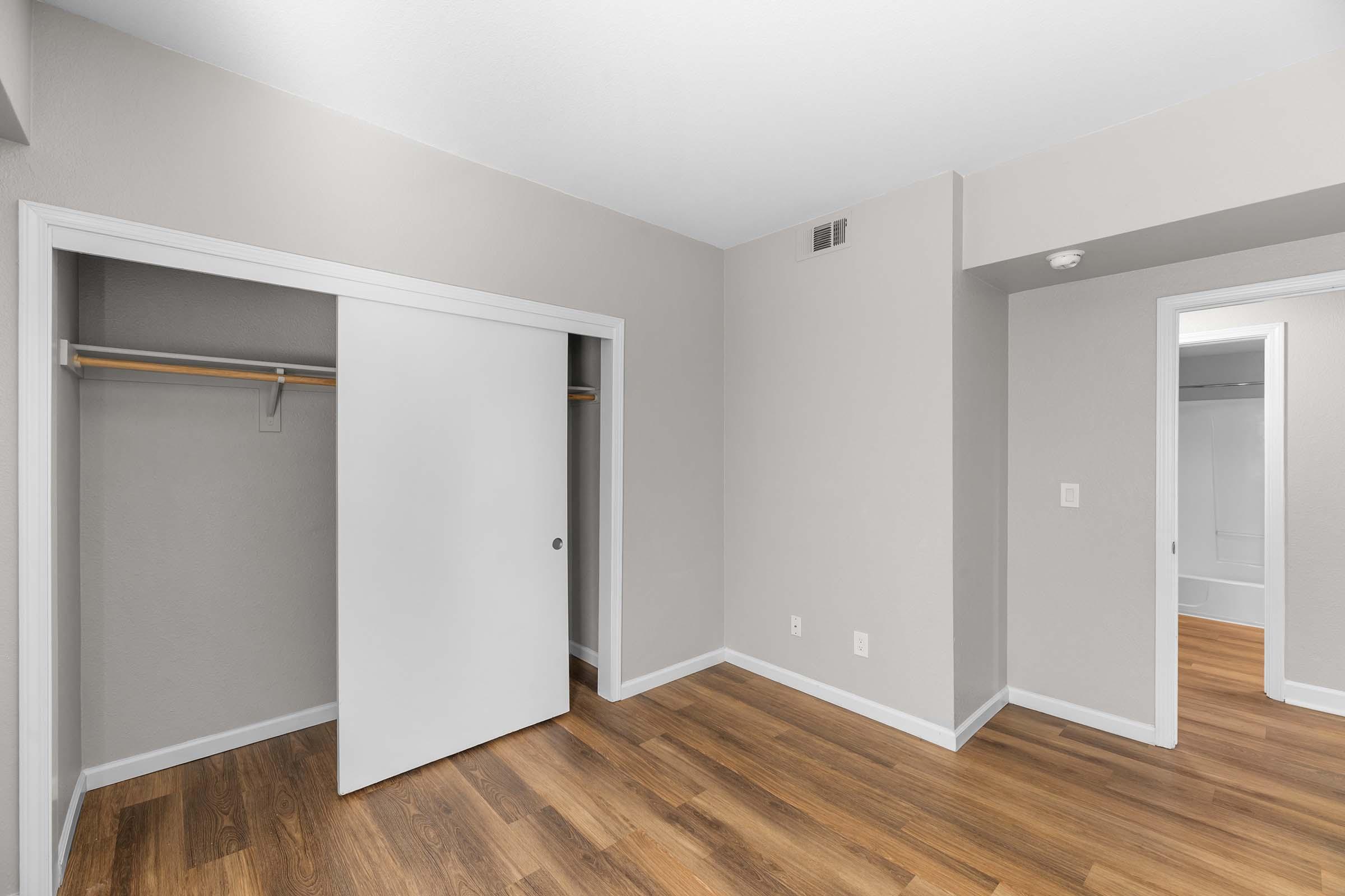 A minimalist bedroom with light gray walls and wooden flooring. A sliding closet door is partially open, revealing a wooden rod for hanging clothes. There is a doorway leading to another room, and the space is well-lit and spacious, creating a clean and modern aesthetic.