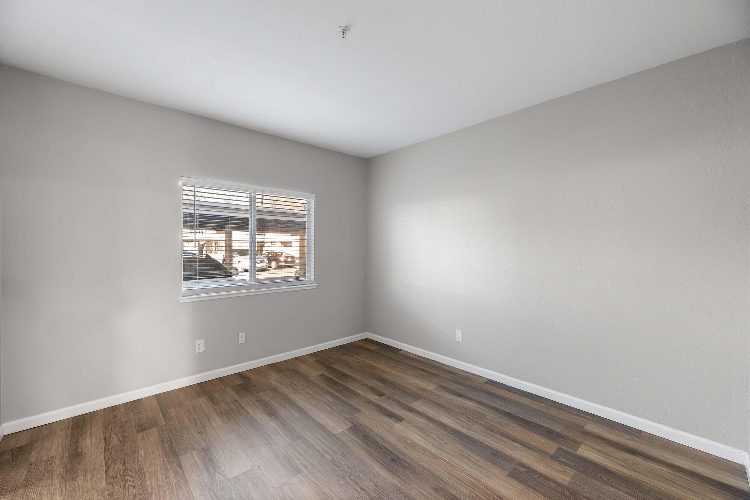 Empty room with light gray walls and wooden flooring. A single window on the left illuminates the space, which has no furniture or decorations. The overall atmosphere is clean and minimalist, showcasing a simple interior design.