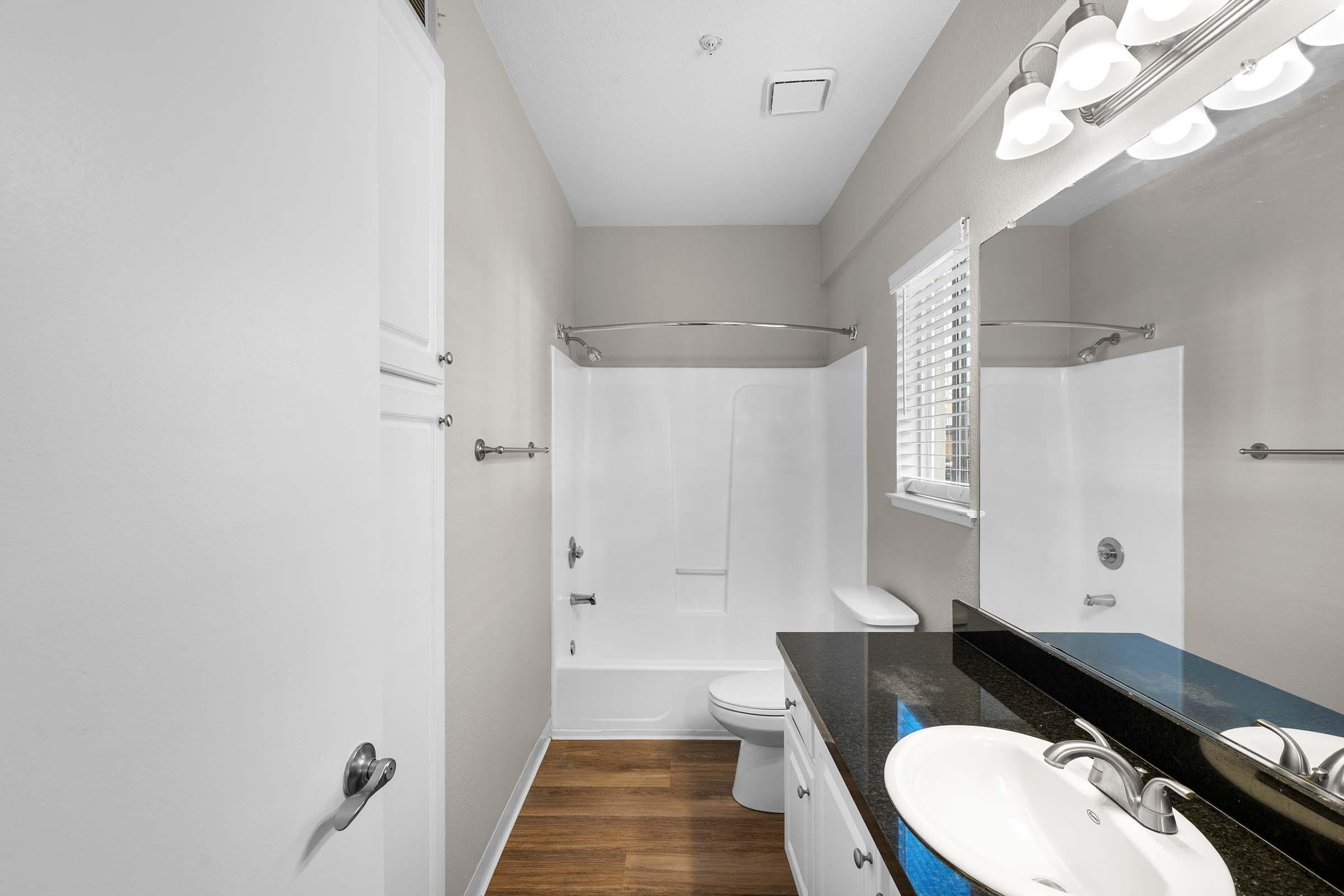 A clean, modern bathroom featuring a white shower/tub combination, a dark granite countertop with a sink, and a mirror above. The walls are painted a neutral color, and there is a window with blinds providing natural light. The flooring is a warm wood tone.