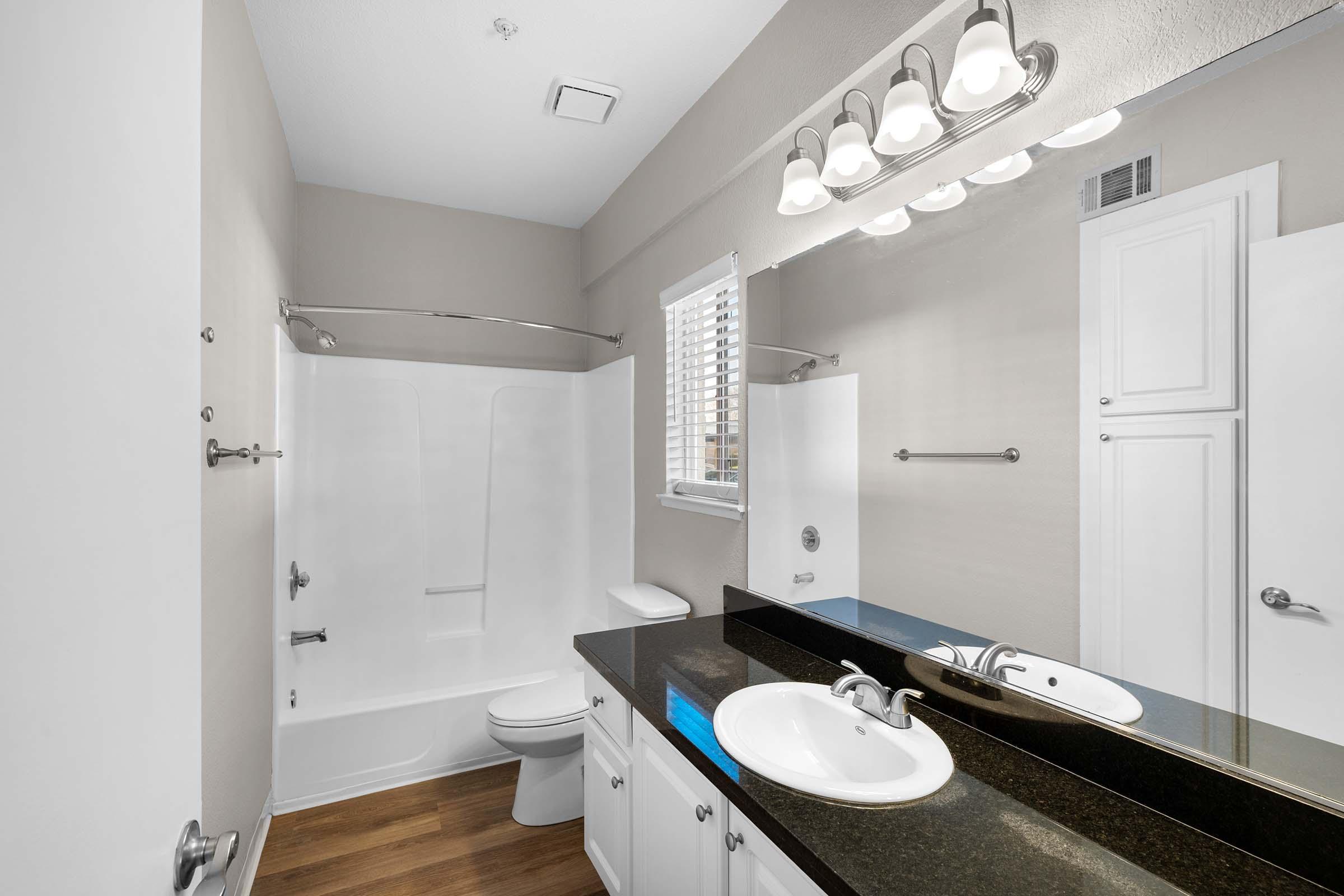 A modern bathroom featuring a white bathtub and shower combination, a toilet, a double sink with a dark countertop, and a large mirror above the sink. Natural light comes through a window with blinds, and the walls are painted in a soft beige color. Wood flooring complements the overall design.