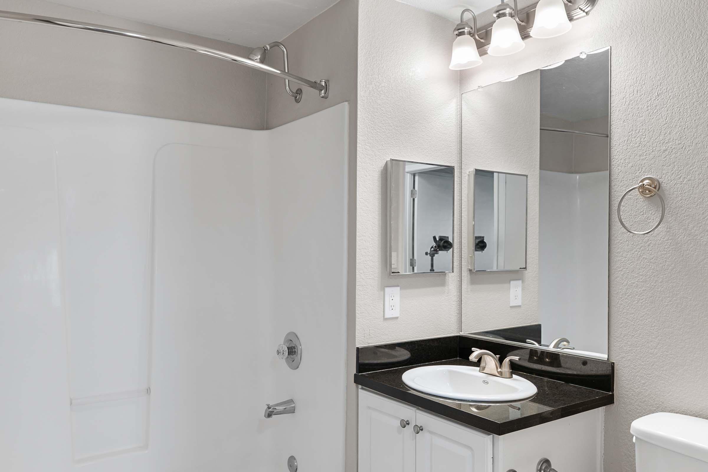 A modern bathroom featuring a white bathtub and shower combo, sleek black countertop with a sink, and a large mirror. The walls are light-colored, and there are two smaller mirrors above the countertop. Contemporary light fixtures are mounted above the mirrors. Clean and minimalist design.