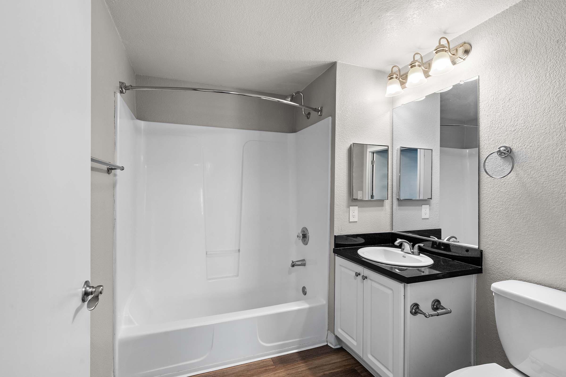A modern bathroom featuring a shower-tub combination with a curved shower rod, a dark countertop with a sink and mirror, white cabinetry, a toilet, and soft lighting. The walls are painted in a light color, and the flooring is wood laminate, creating a clean and contemporary look.