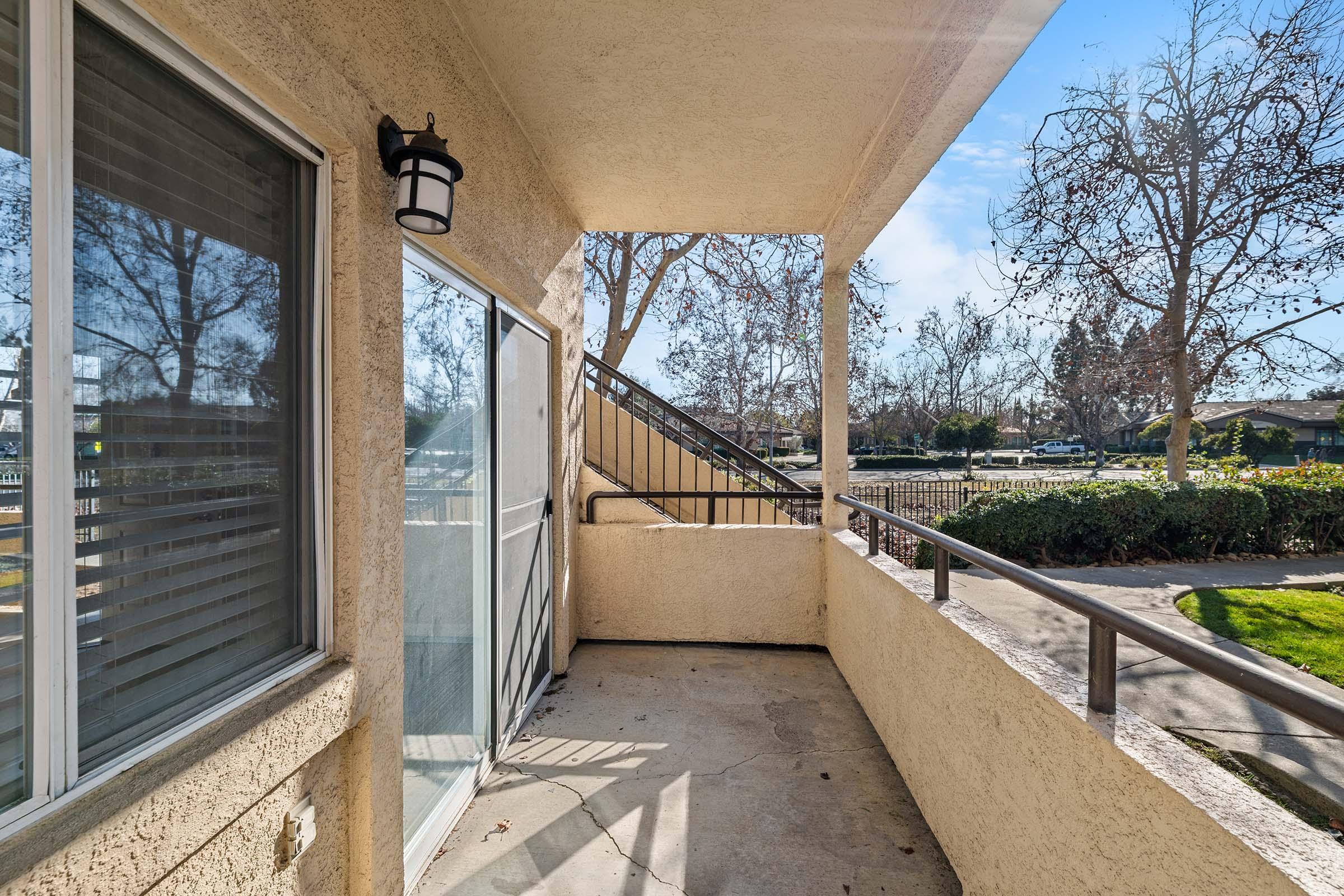 A balcony view from an apartment featuring a concrete surface, with a stairway leading down and trees visible in the background. Natural light illuminates the area, highlighting the beige exterior walls.