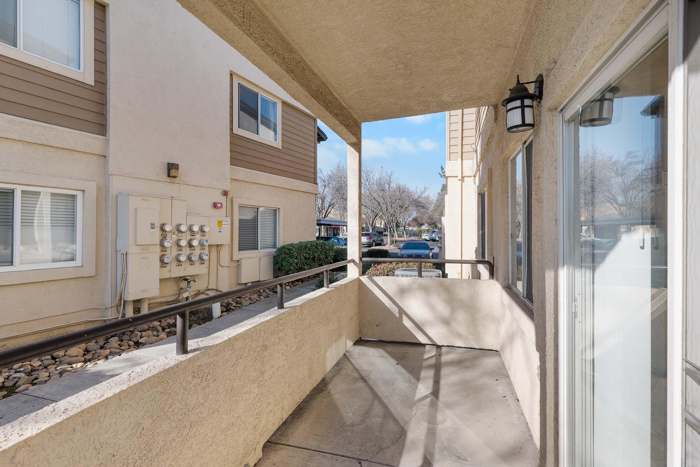 A view from a covered balcony showing the exterior of an apartment building. The balcony features a railing and has smooth flooring. In the background, another building is visible with utility meters, greenery, and trees, under a clear blue sky. The scene is well-lit and inviting.